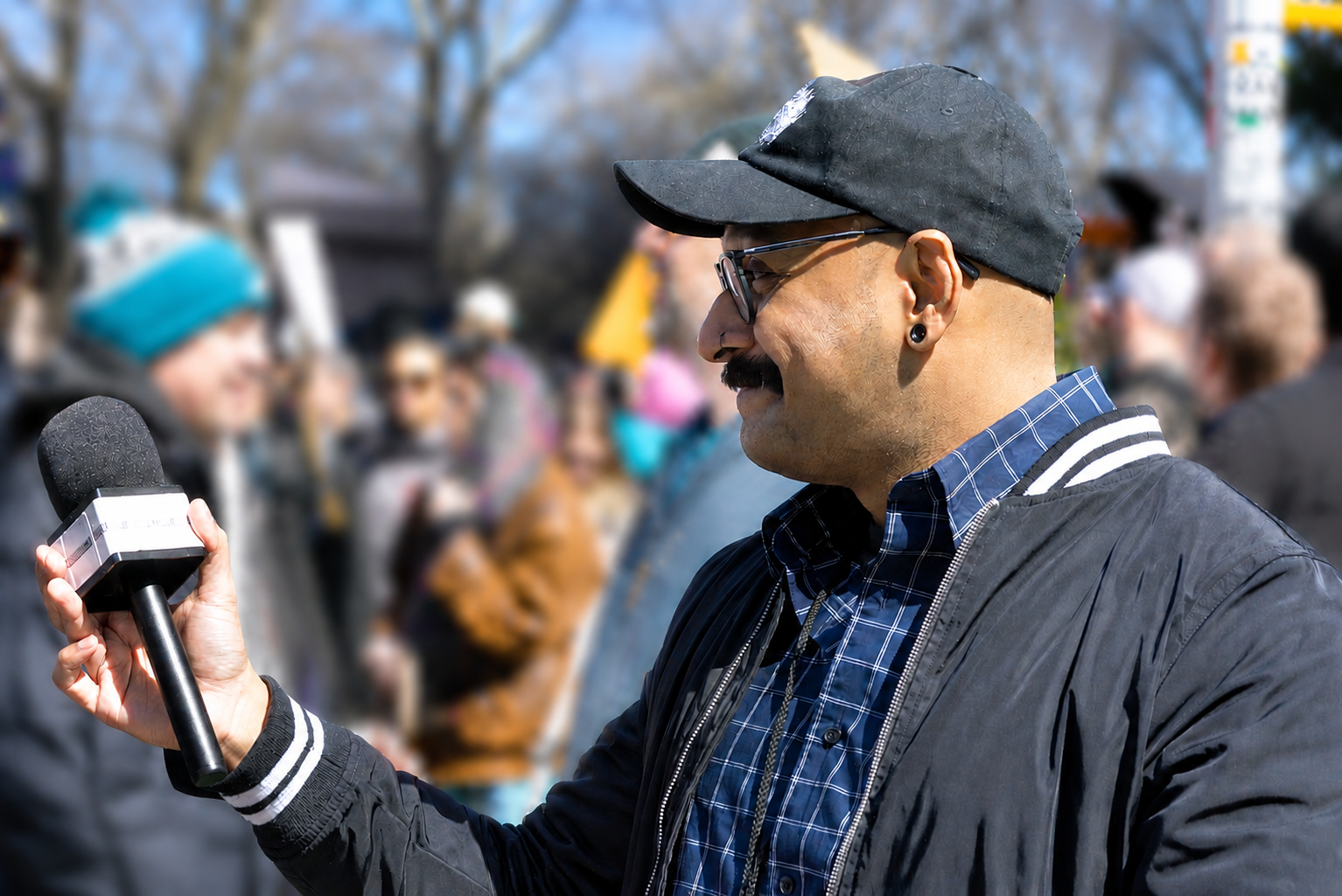 Andy Sahadeo holding a microphone during No Kings NYC with a crowd in the background, wearing a black cap, glasses, and a black jacket. Coverage for No Bullshit.