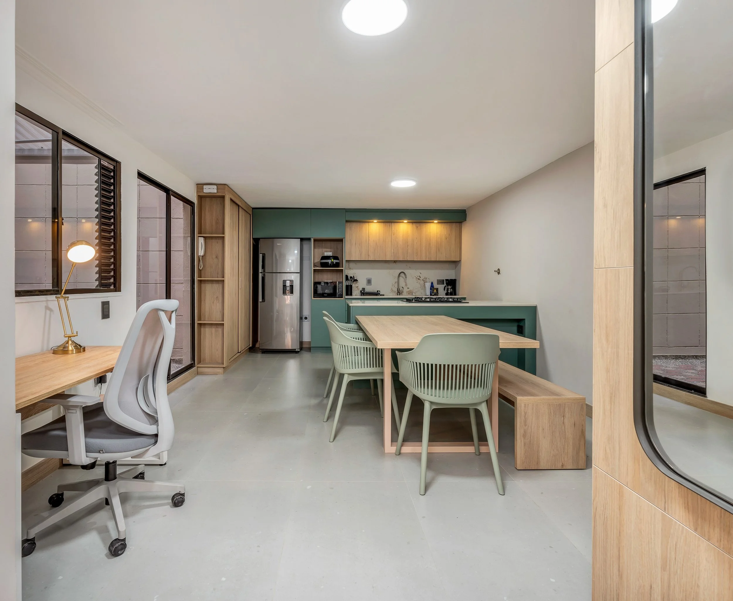 Vista de una cocina moderna y luminosa con gabinetes en tonos verde y madera, una mesa de madera y sillas blancas, electrodomésticos metálicos y ventanas grandes con persianas de listones.
