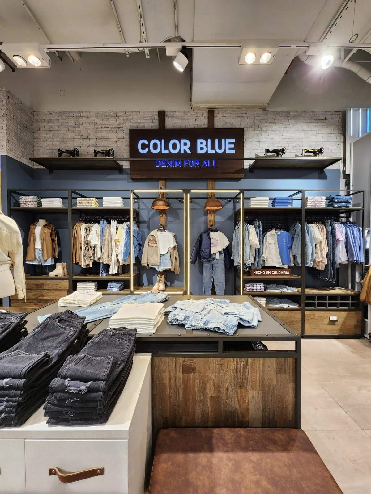 Interior de una tienda de ropa con estanterías llenas de prendas de mezclilla y camisetas, y un cartel que dice 'COLOR BLUE' y 'DENIM FOR ALL'.