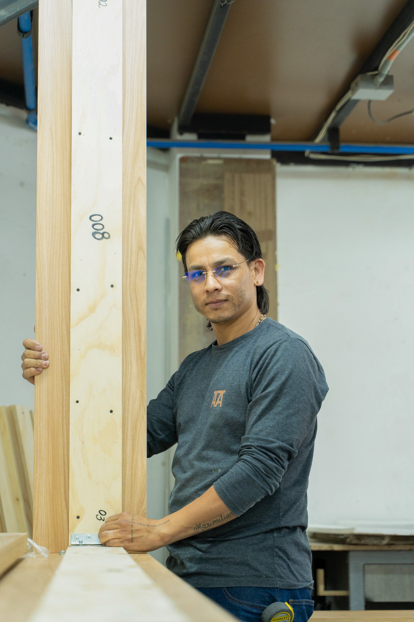Hombre con gafas de sol azules sosteniendo una tabla de madera en una carpintería.