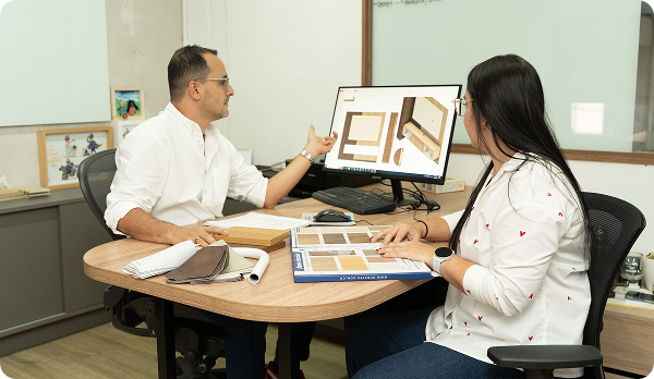 Dos personas en una oficina discuten ideas frente a una computadora y muestras de diseños de muebles en la mesa.