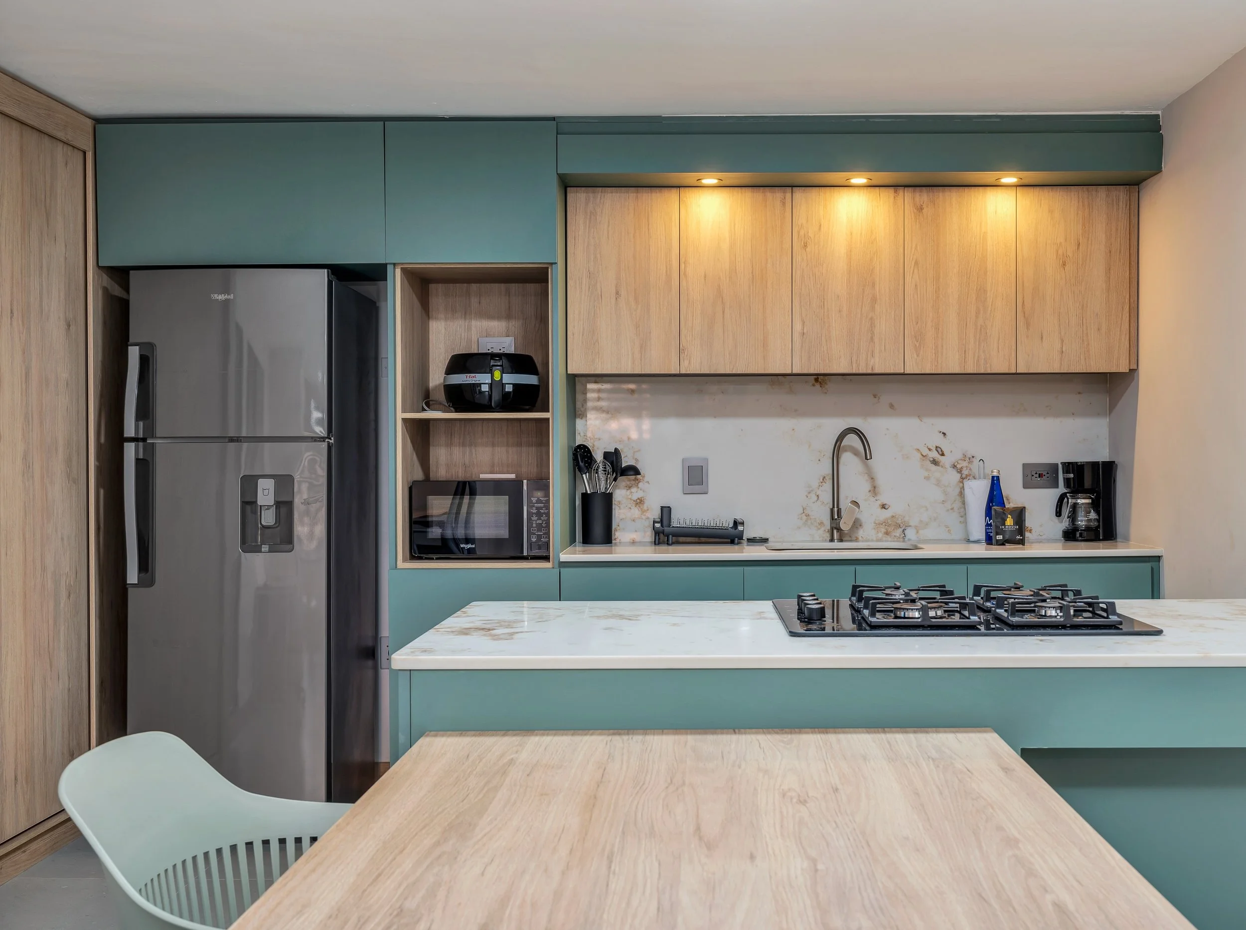 Cocina moderna con nevera, microondas, gabinetes de madera y encimera de mármol, vista desde la mesa en primer plano.