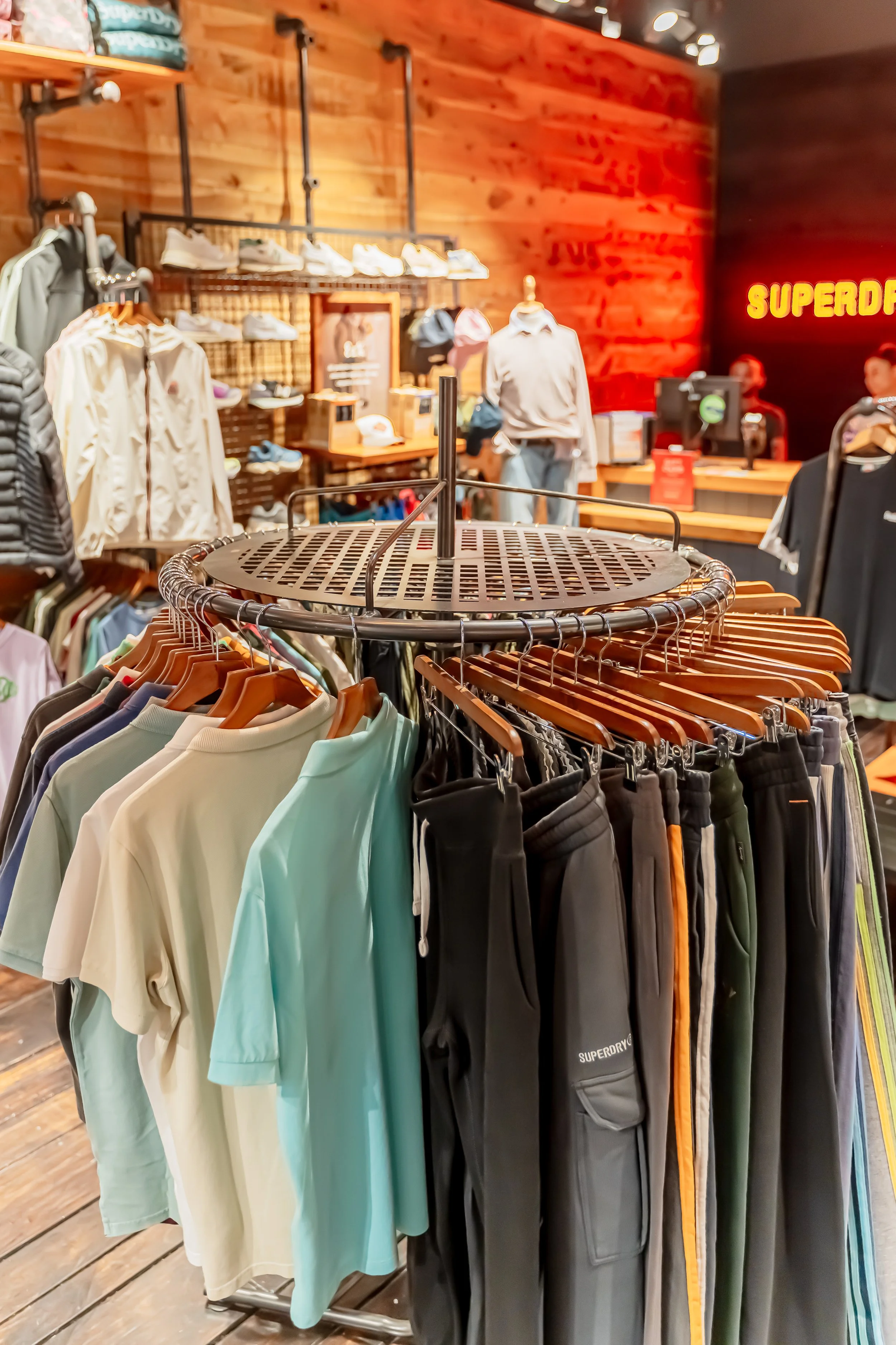 Prenda de ropa en un perchero con varias camisetas y pantalones en una tienda de ropa, fondo de paredes de madera y estanterías con calzado y accesorios, luces en el techo y una señal de neón que dice 'Superdry'.