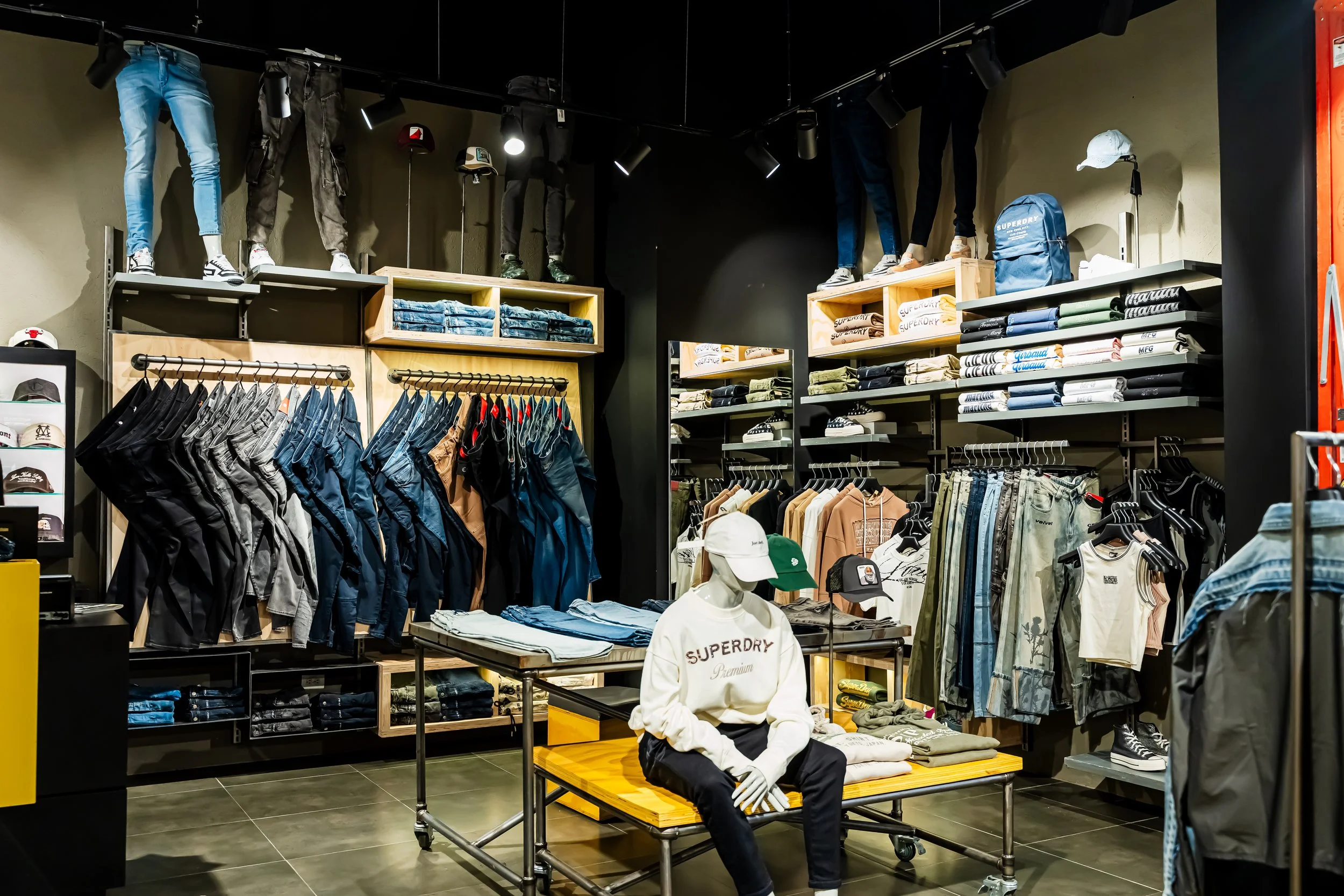 Vista interior de una tienda de ropa con maniquíes con pantalones y accesorios, estantes con ropa doblada y prendas colgadas en perchas, y sombreros.