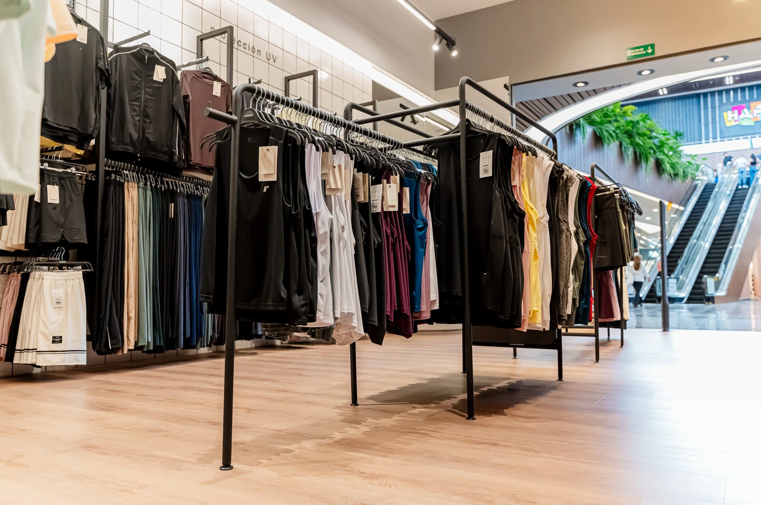 Ropa deportiva y casual colgada en percheros en una tienda, con escaleras mecánicas y letrero en el fondo.