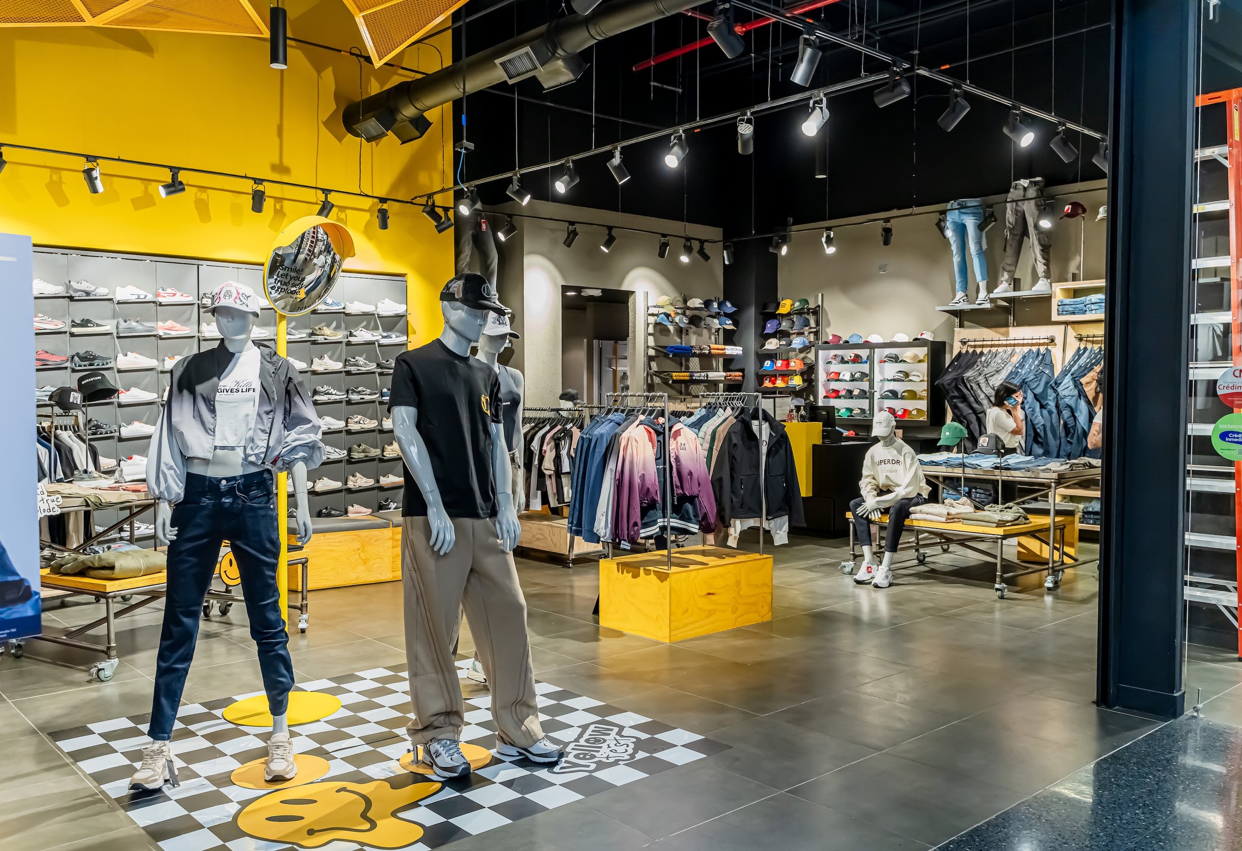 Vista de una tienda de ropa y calzado con maniquíes de figuras humanas vestidas con ropa deportiva y casual, gafas, y sombreros. Hay estantes con zapatos y gorras, con un fondo de paredes pintadas en amarillo y negro, con iluminación de focos en el techo.