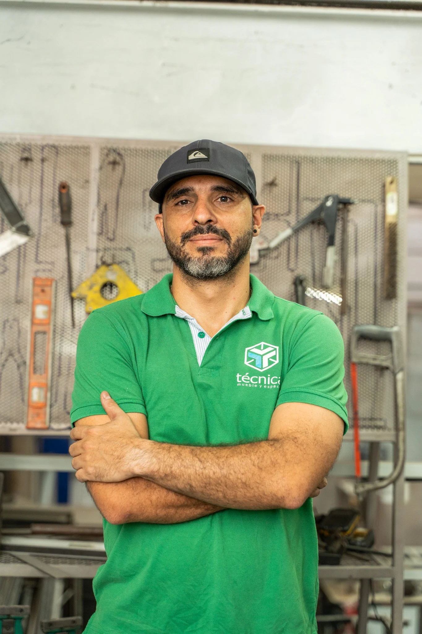 Hombre con barba, camiseta verde y gorra, posando con los brazos cruzados en taller de herramientas.