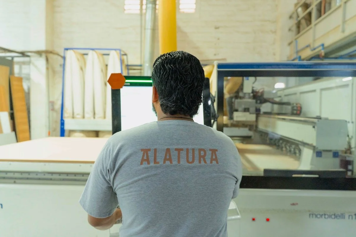 Persona con camiseta gris que tiene la palabra 'ALTURA' en la parte trasera, trabajando en una máquina en un taller o fábrica.