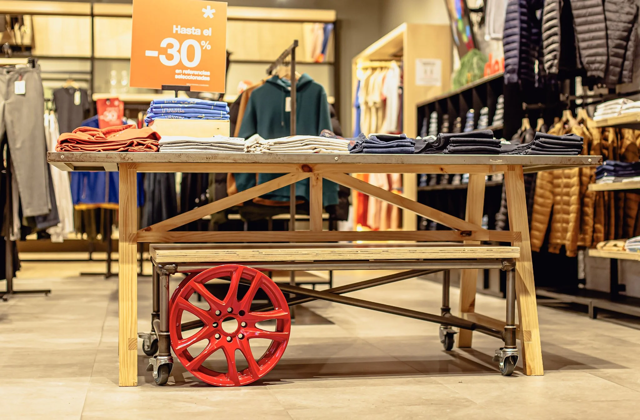 Mesa de madera con prendas de ropa dobladas y cartel naranja con promoción de hasta 30% de descuento en rebajas, fondo de tiendas con ropa en exhibición.