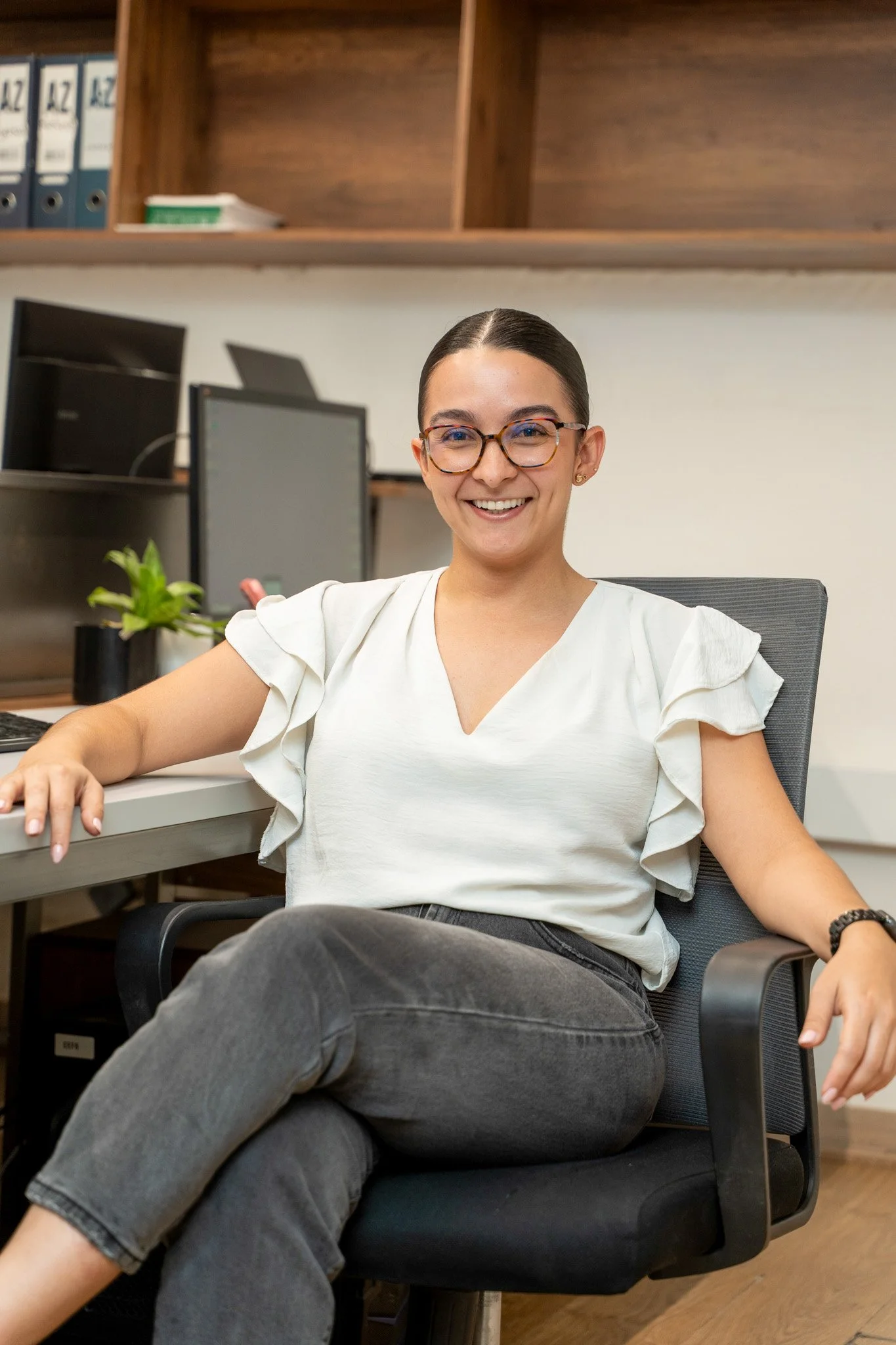 Mujer joven con gafas de marco grande sonriendo, sentada en una oficina con computadora y estanterías de libros y archivos