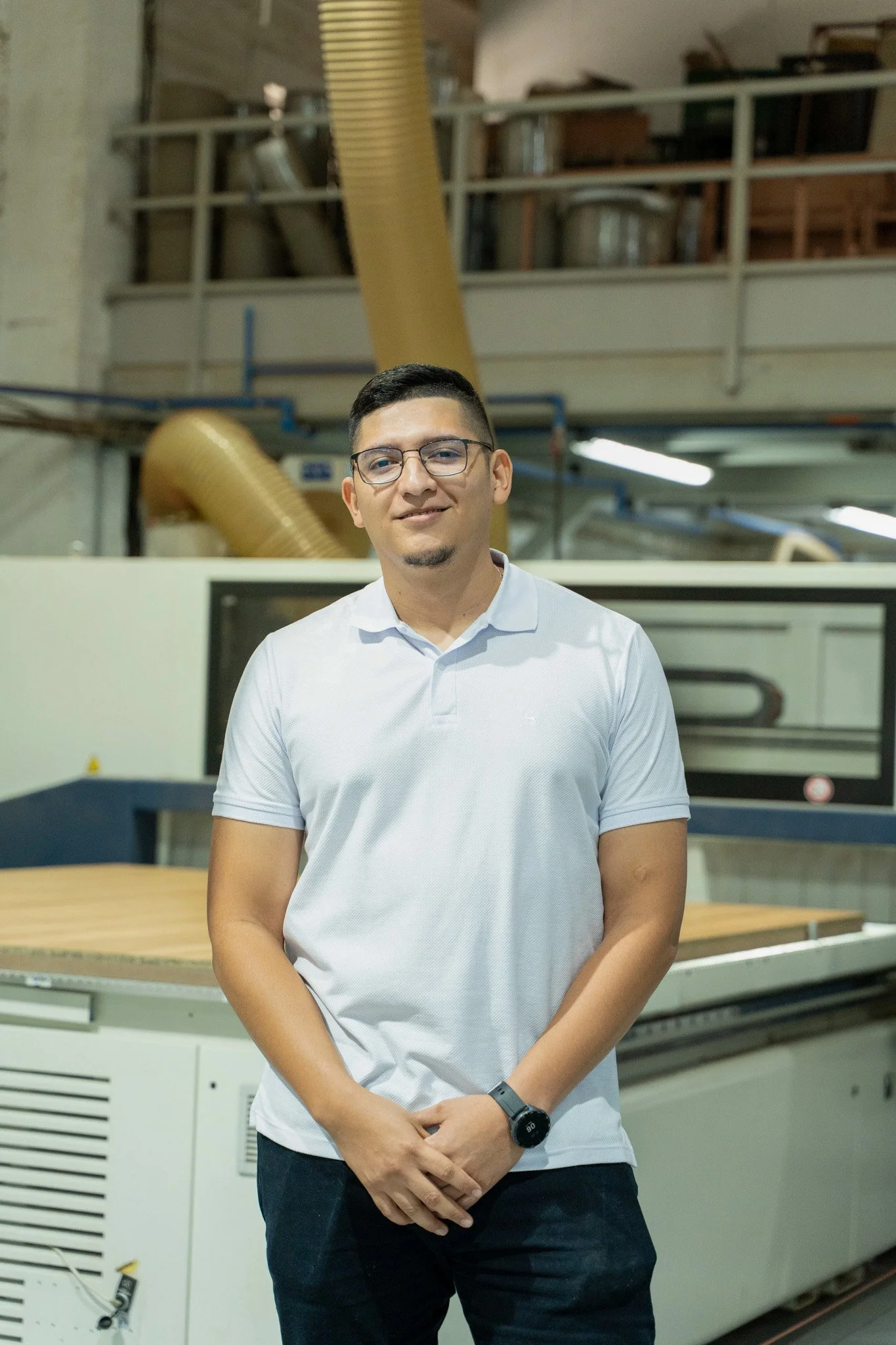 Hombre con gafas y camiseta blanca de pie en un taller, con maquinaria y tubos de ventilación en el fondo.