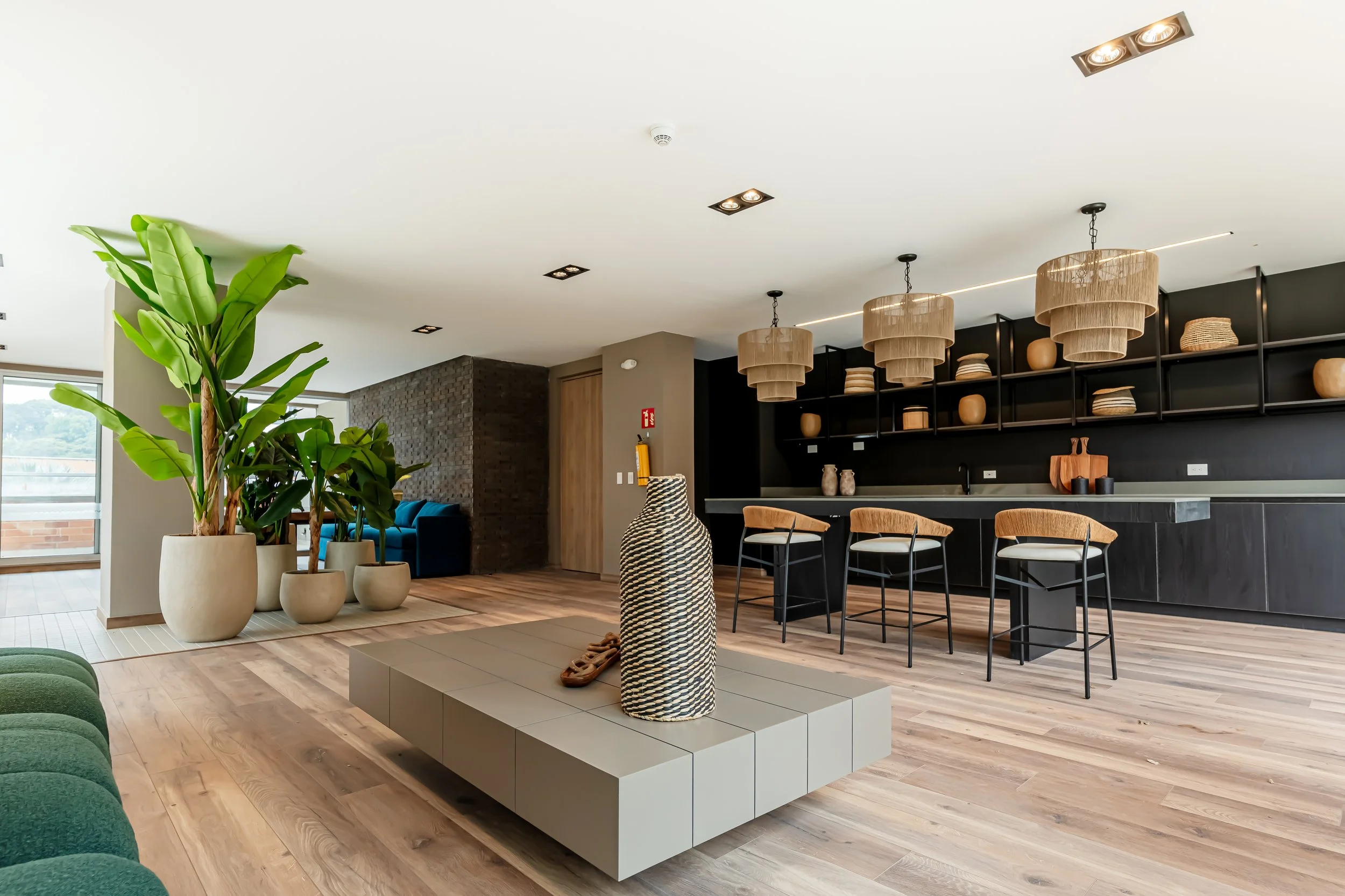 Interiores modernos con plantas verdes, mesas decorativas, lámparas de mimbre, sillas de madera y pared con estantes de cerámicas y cestas, en ambiente de habitación o sala de estar.