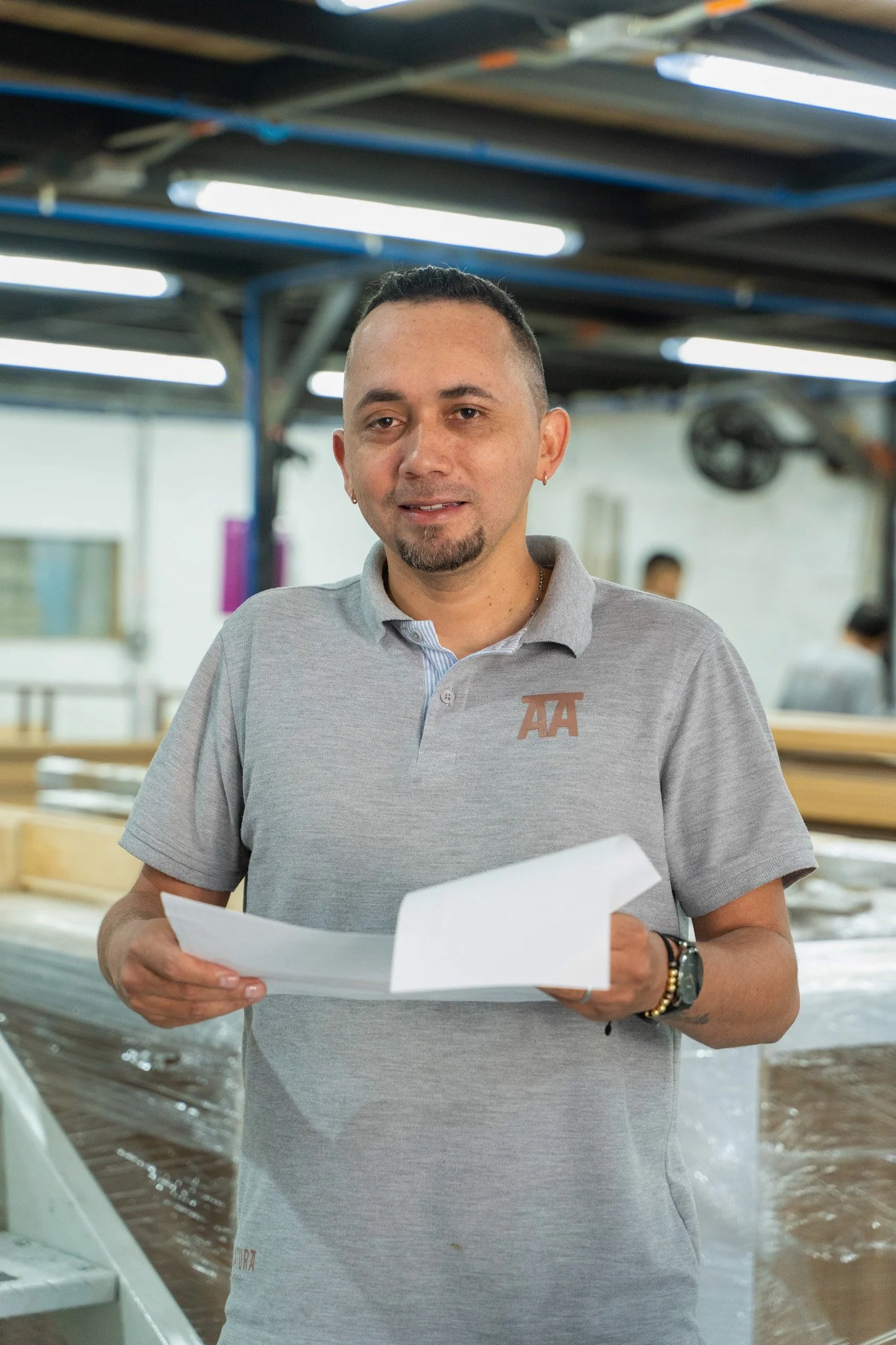 Un hombre con gafas y con barba, vestido con una camiseta gris y con peinado de corte militar, sosteniendo documentos en una mano, en un entorno industrial o de fábrica, con maquinaria y estructuras metálicas en el fondo.