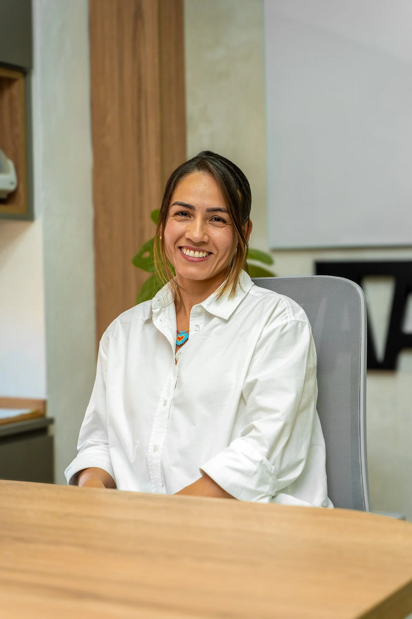 Retrato de una mujer sonriendo, sentada en una sala de oficina con fondo de cortinas y mobiliario.
