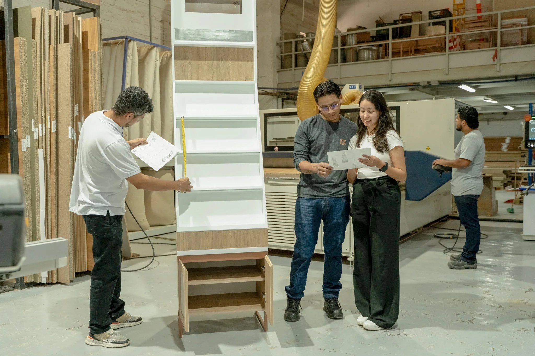 Personas en una fábrica de madera revisando planos y mostrando una estantería de madera, con maquinaria y productos de madera en el fondo.