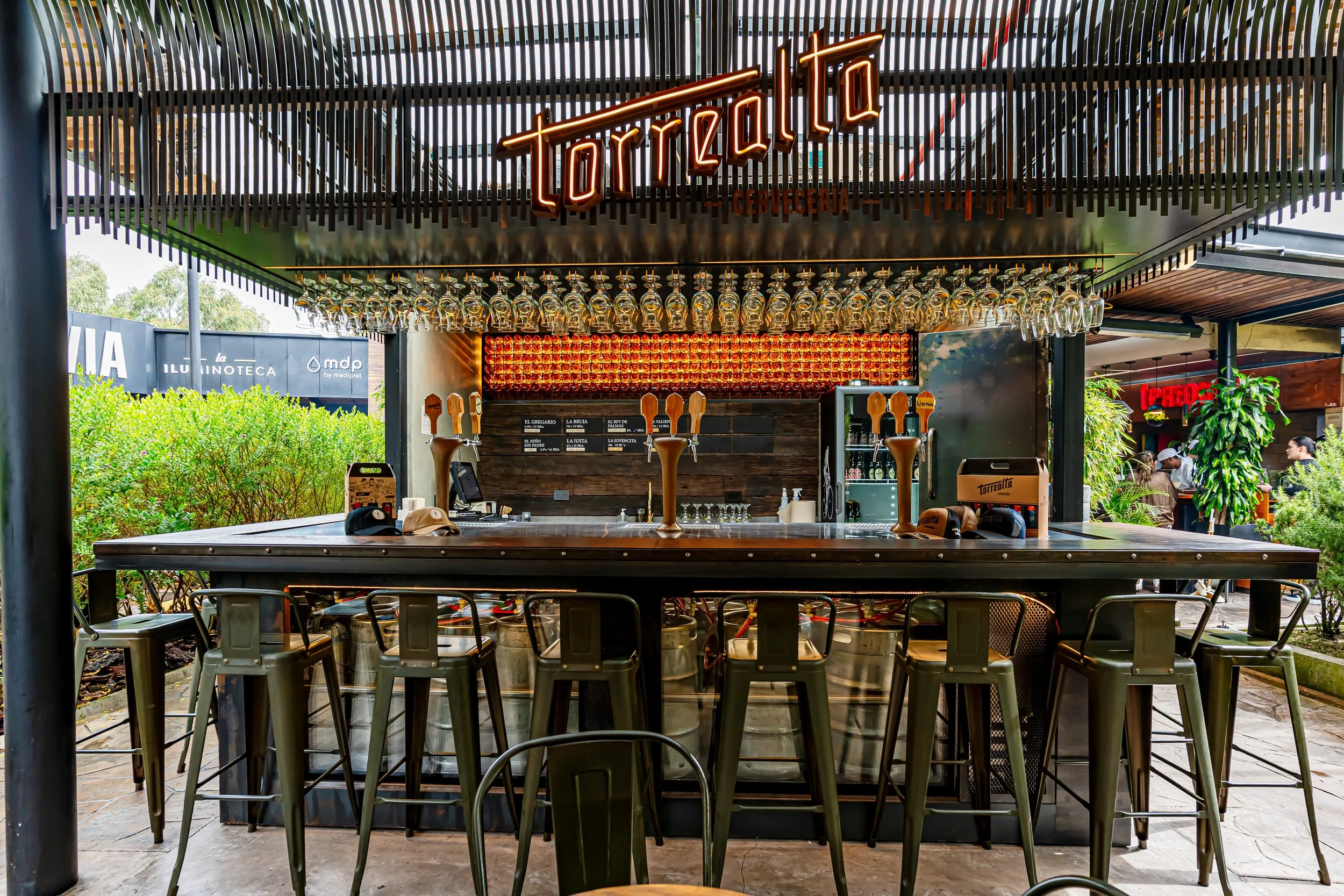 Bar de cerveza con letras de neón que dice 'Tarralio', con copas de vidrio colgando, en un ambiente interior y exterior con vegetación y personas en el fondo.