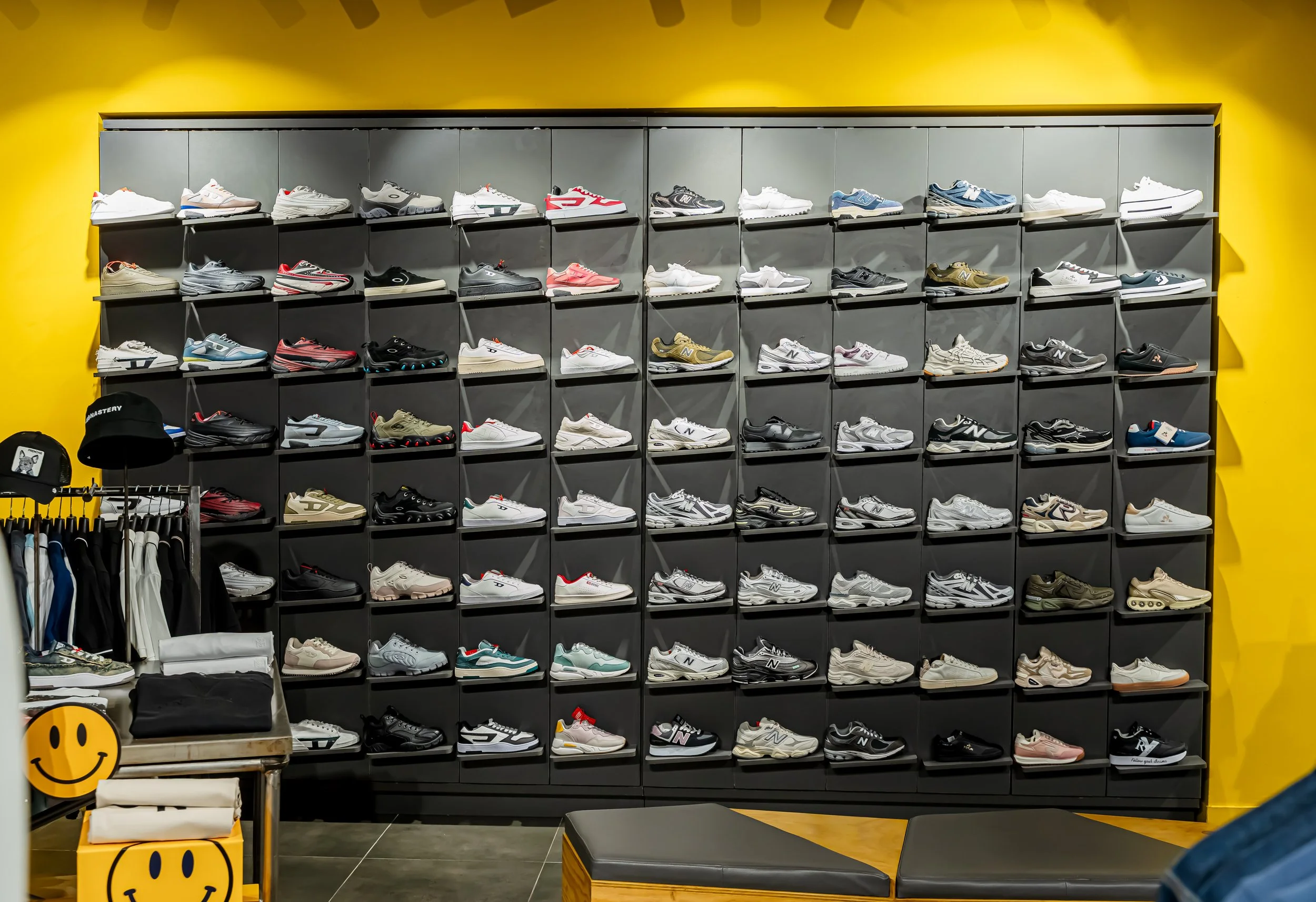 Estantería gris con múltiples pares de zapatos deportivos de diferentes estilos y colores, sobre un fondo de pared amarilla en una tienda.