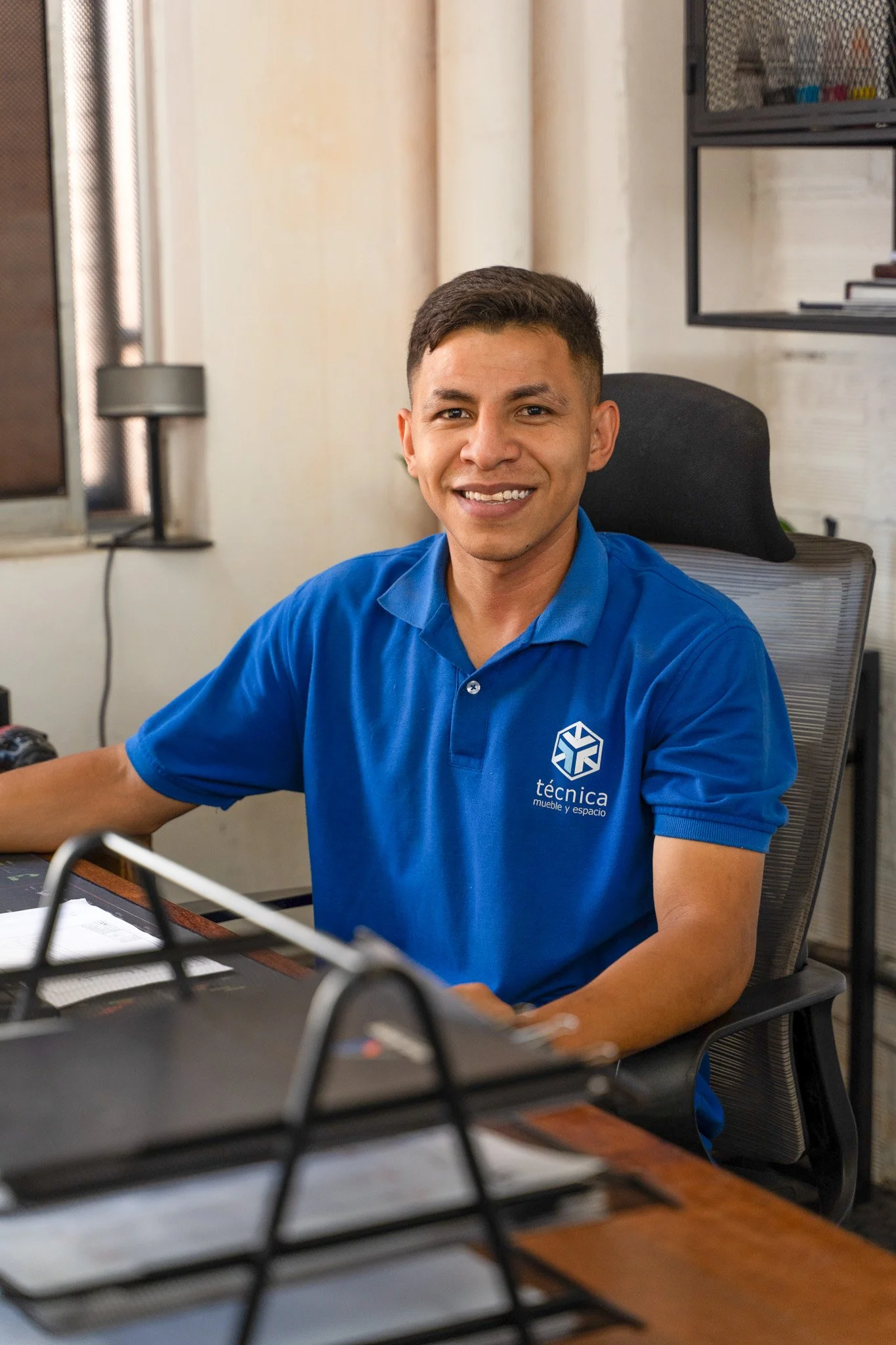 Joven sonriendo sentado en una oficina, lleva una camisa azul con un logotipo y la palabra 'técnica' en ella. Está frente a una mesa con objetos y detrás de una silla ergonómica.