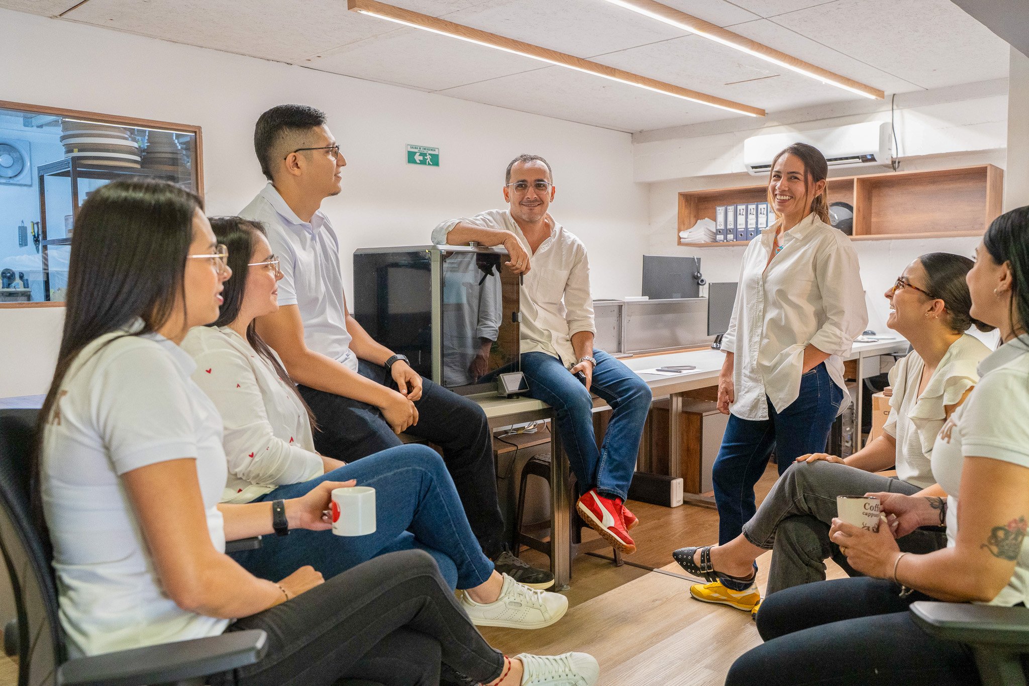 Grupo de personas en una reunión en una oficina, sentadas en círculo, escuchando y participando con sonrisas. Algunas toman café y hay archivos en estanterías.
