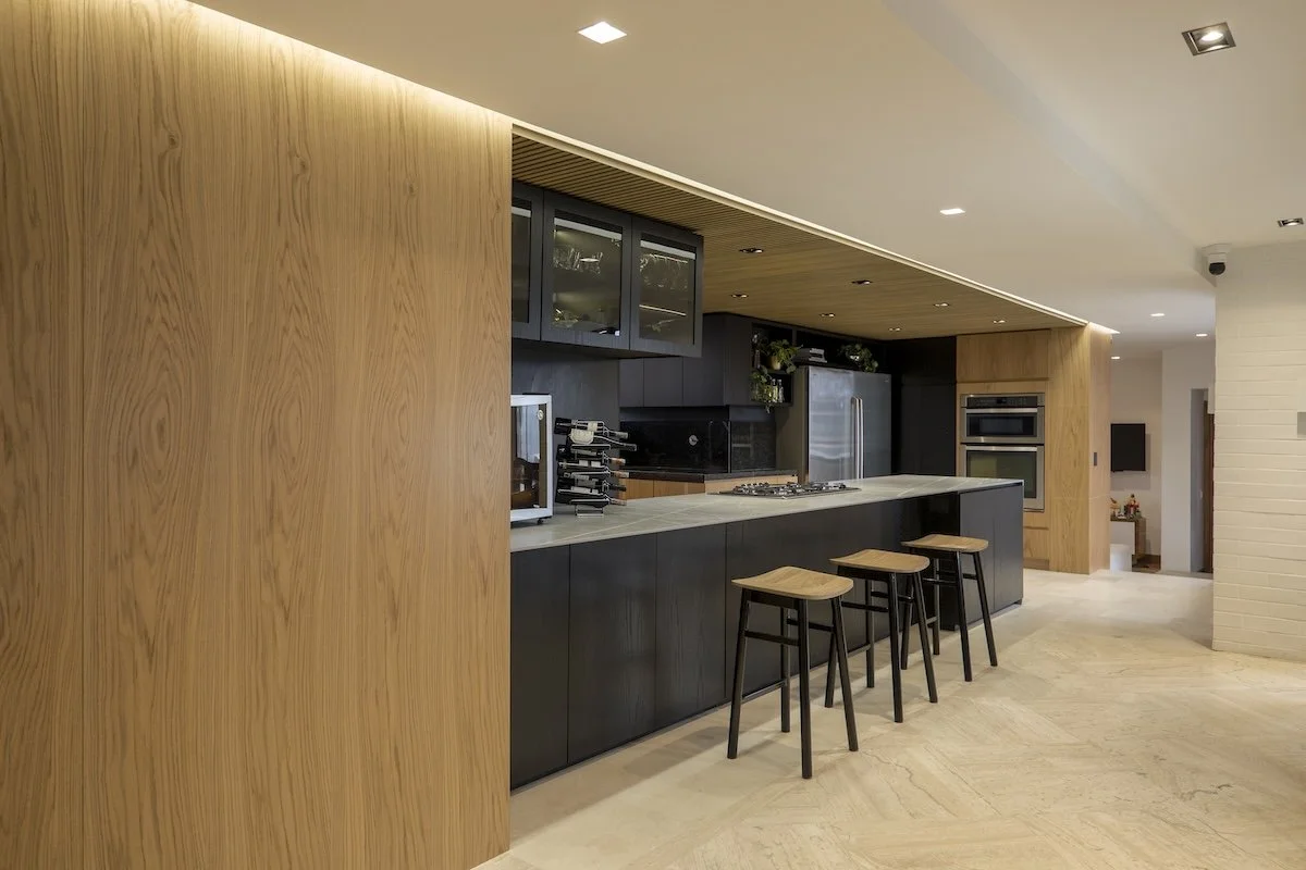 Cocina moderna con paredes de madera, gabinetes oscuros y cinco bancos de madera junto a una isla de cocina con estufa y electrodomésticos de acero inoxidable.