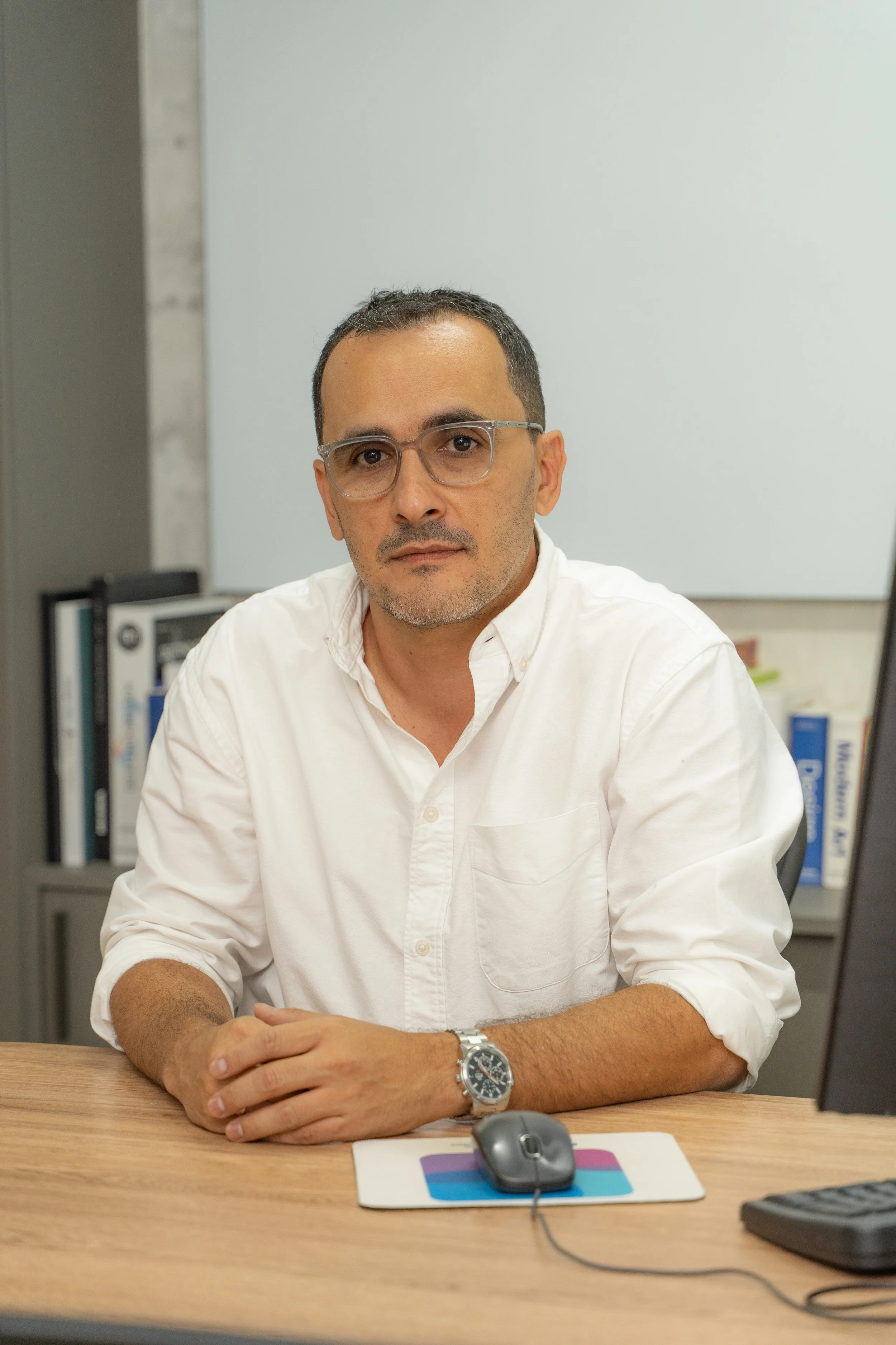 Hombre con gafas y camisa blanca sentado en una mesa de oficina, viendo a la cámara, con computadora y ratón en la mesa.