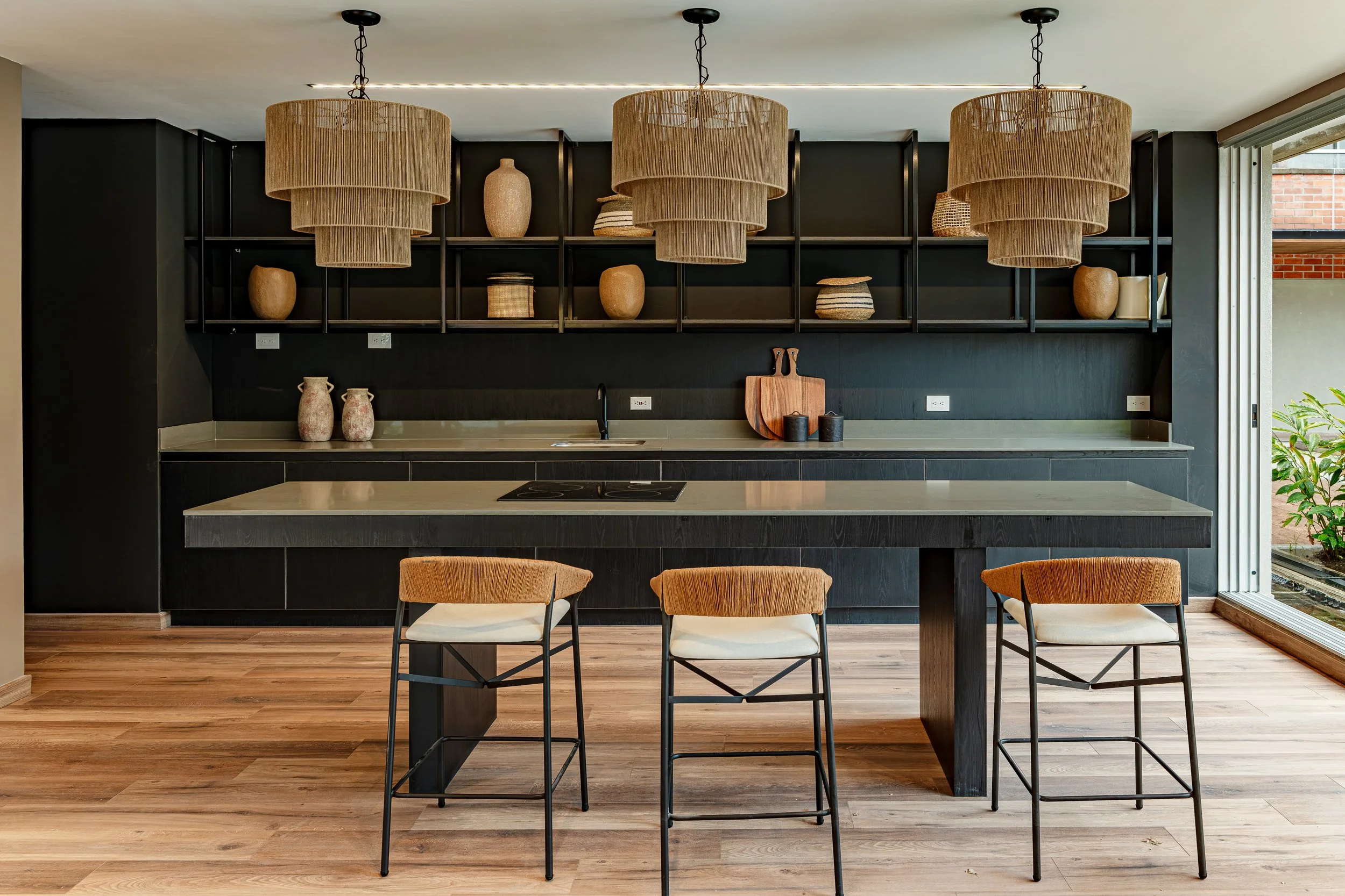 Cocina moderna con mesa grande, sillas de diseño y estantería negra en la pared con jarrones y cestos de barro, iluminada con lámparas de mimbre colgantes.