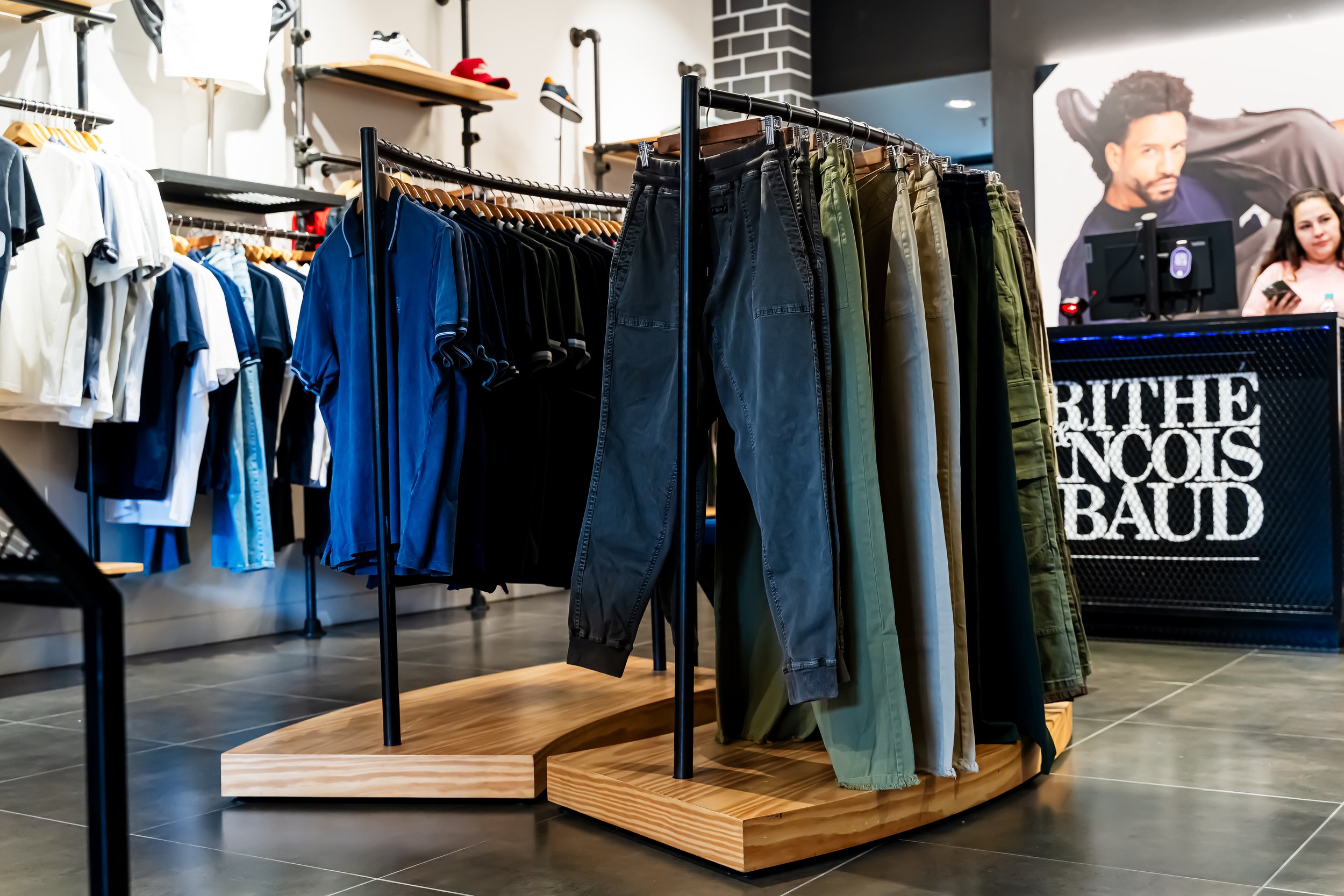 Ropa de diferentes colores y estilos en exhibición en una tienda de moda, con una mujer en la pantalla gigante de fondo.
