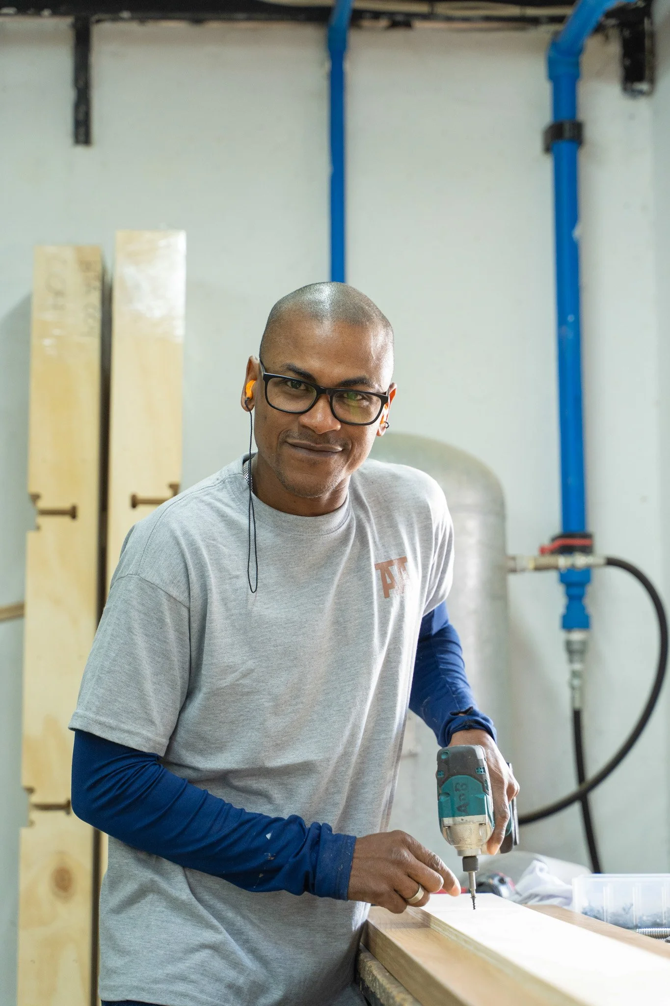 Hombre con gafas y auriculares trabajando con una herramienta eléctrica en un taller, con tuberías azules en la pared trasera.