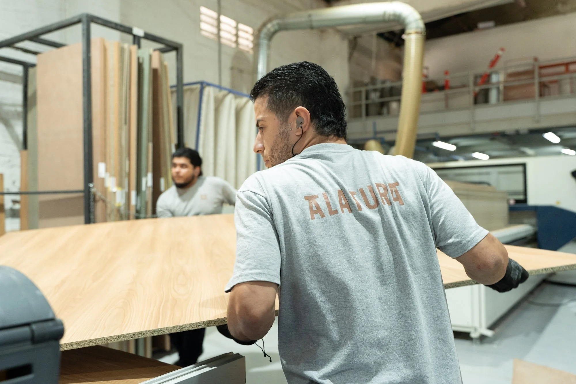 Joven hombre con audífonos en un almacén de madera y tableros de laminado, sosteniendo una pieza de madera en una mano.
