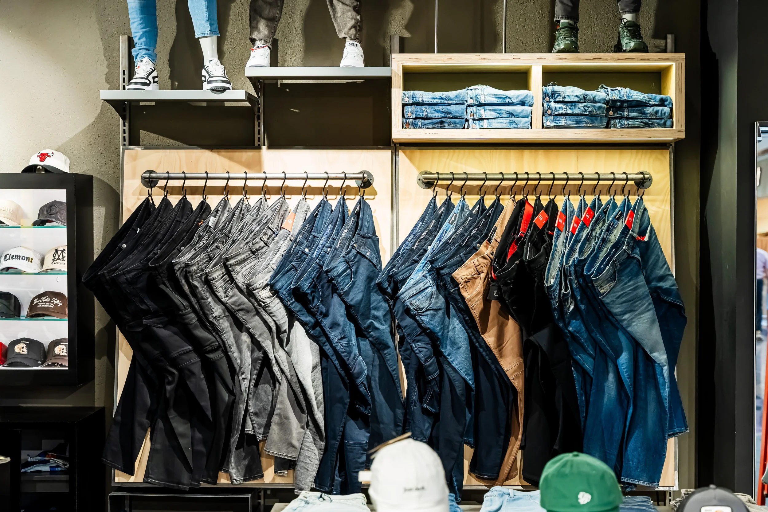 Varios pantalones y shorts de mezclilla colgados en perchas en una tienda de ropa. En la parte superior hay estantes con pantalones doblados y calzado. A la izquierda, una sección con gorras y sombreros.