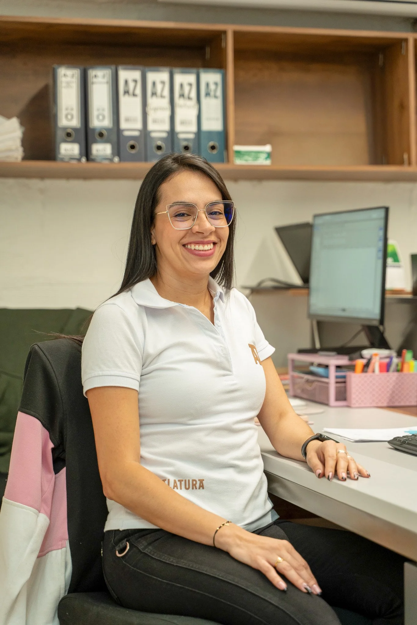 Una mujer sonriendo, sentada en una oficina, lleva gafas, camisa blanca y pantalones oscuros. Tiene cabello largo oscuro y usa un reloj negro en la muñeca derecha. Tras ella hay un escritorio con papeles, una computadora y organizadores de oficina en tonos rosa y azul. Al fondo, estanterías de madera con archivadores y papeles.