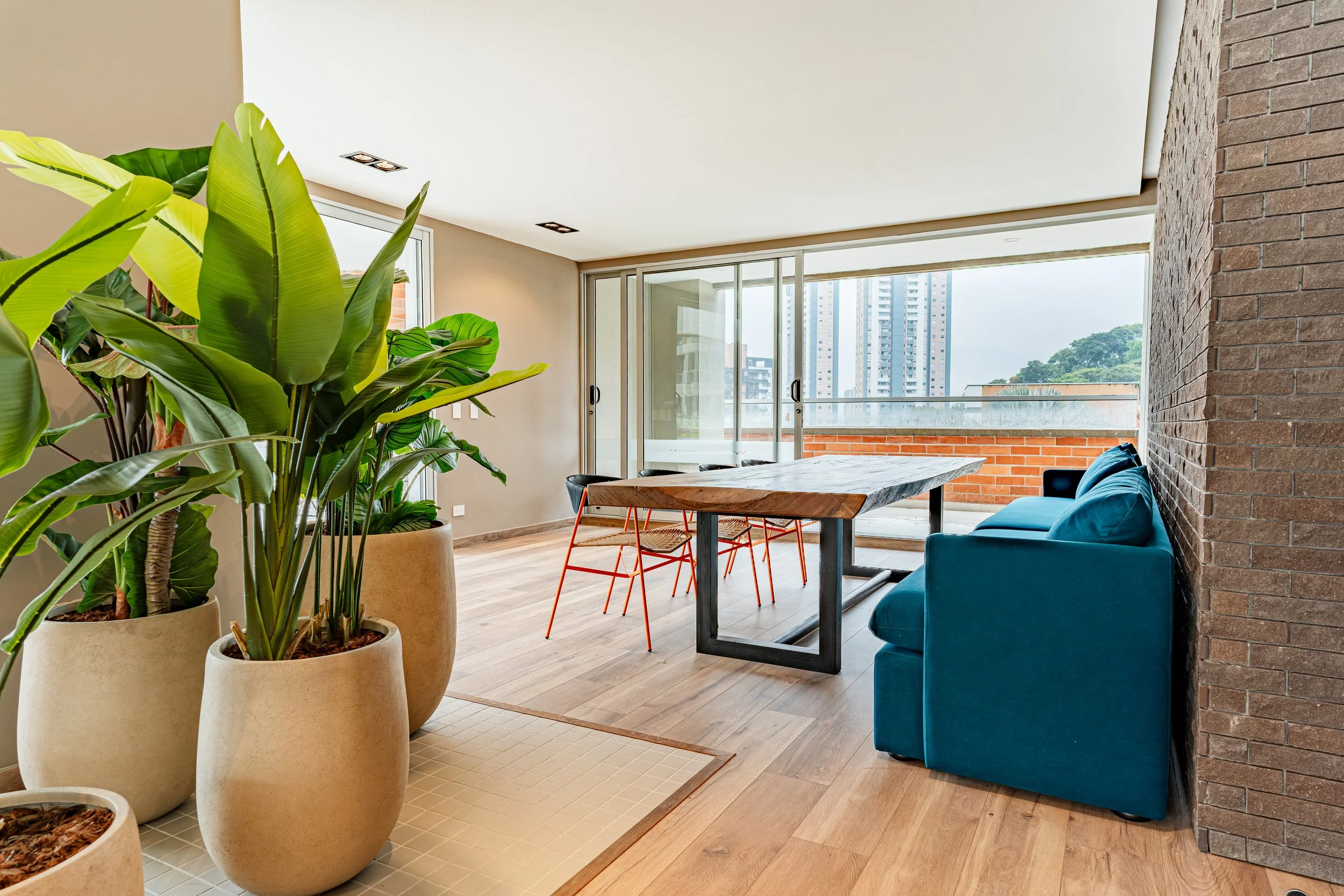 Interior de un apartamento con plantas verdes en macetas grandes, una mesa de madera, sillas de color naranja, un sofá azul y un ventanal panorámico que muestra edificios altos y árboles.