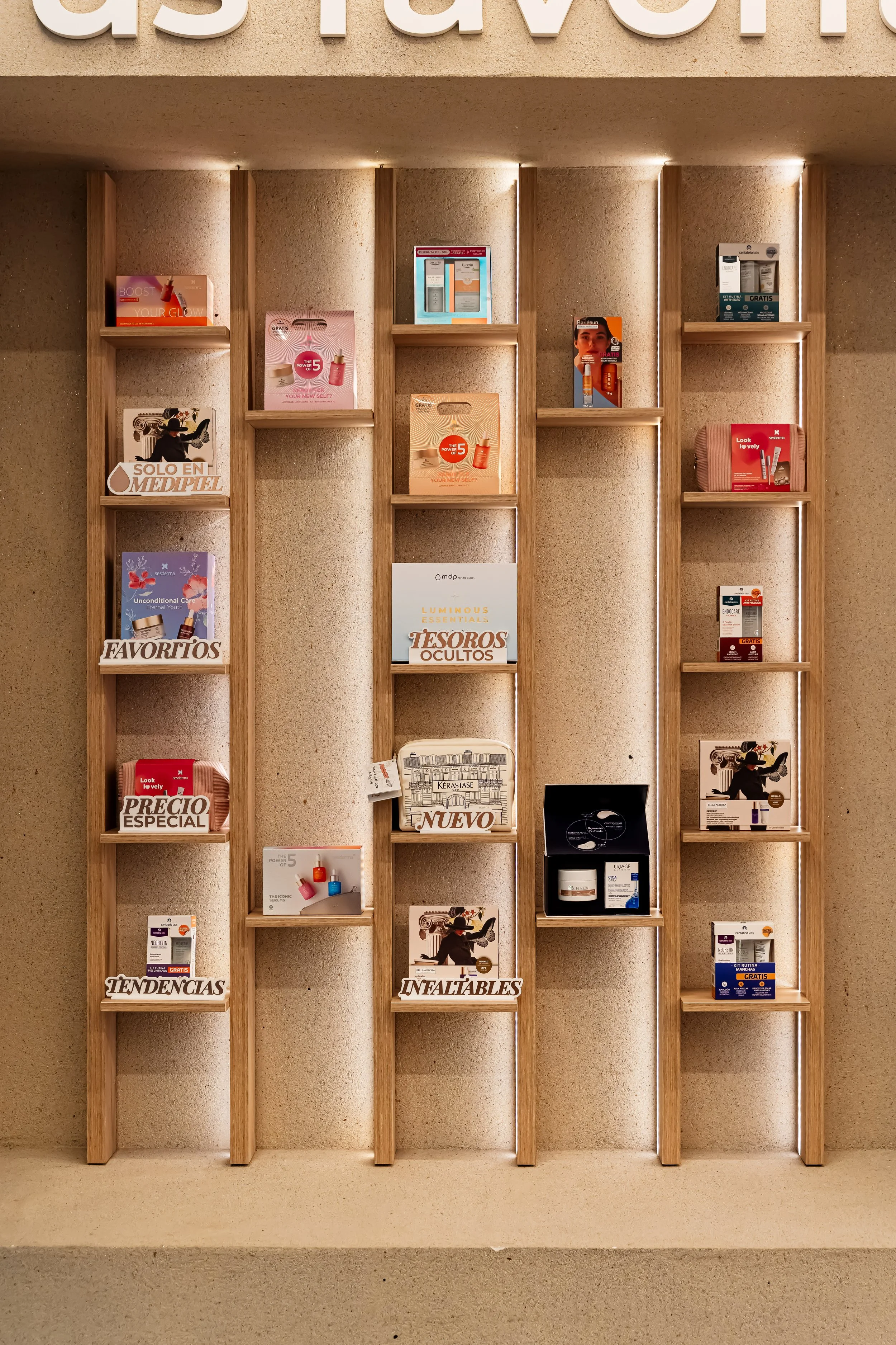 Estantería de madera con varias revistas y promociones de productos de belleza, cuidado de la piel y medicina estética, organizadas en diferentes secciones con etiquetas en español como 'Favoritos', 'Tendencias', 'Infaltables', 'Novedad', 'Tesoros Ocultos', 'Precio Especial', en un entorno interior de pared beige.