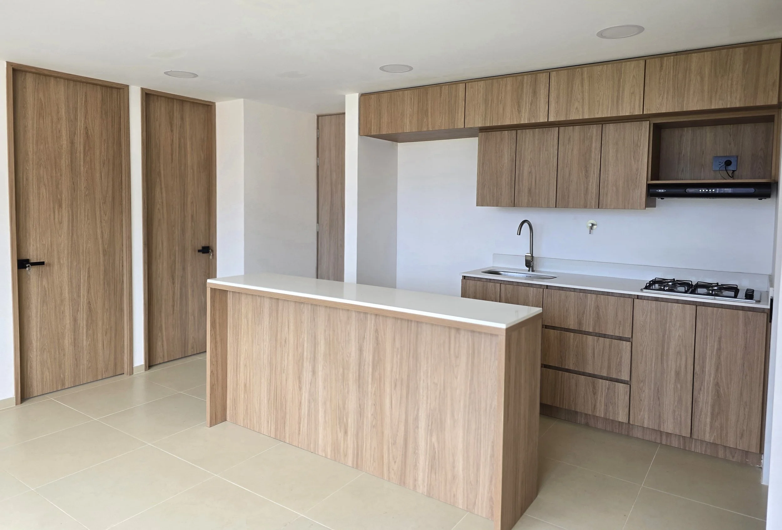 Cocina moderna con gabinetes de madera y encimera blanca, fregadero, estufa de gas, campana extractora y enchufe en la pared.