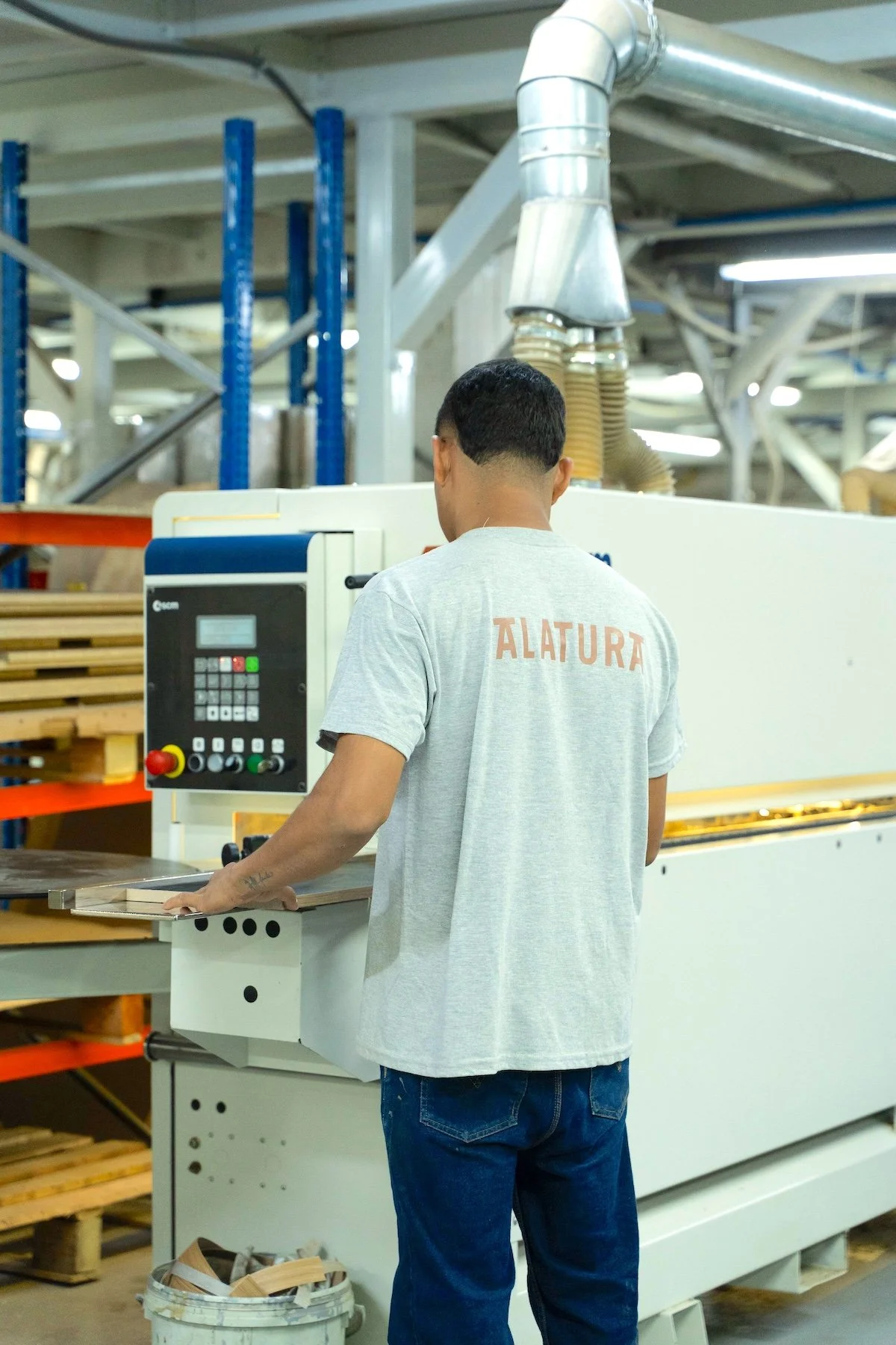Persona trabajando en una máquina en un taller de madera, llevando una camiseta gris con la palabra 'ALTURA' en la espalda.