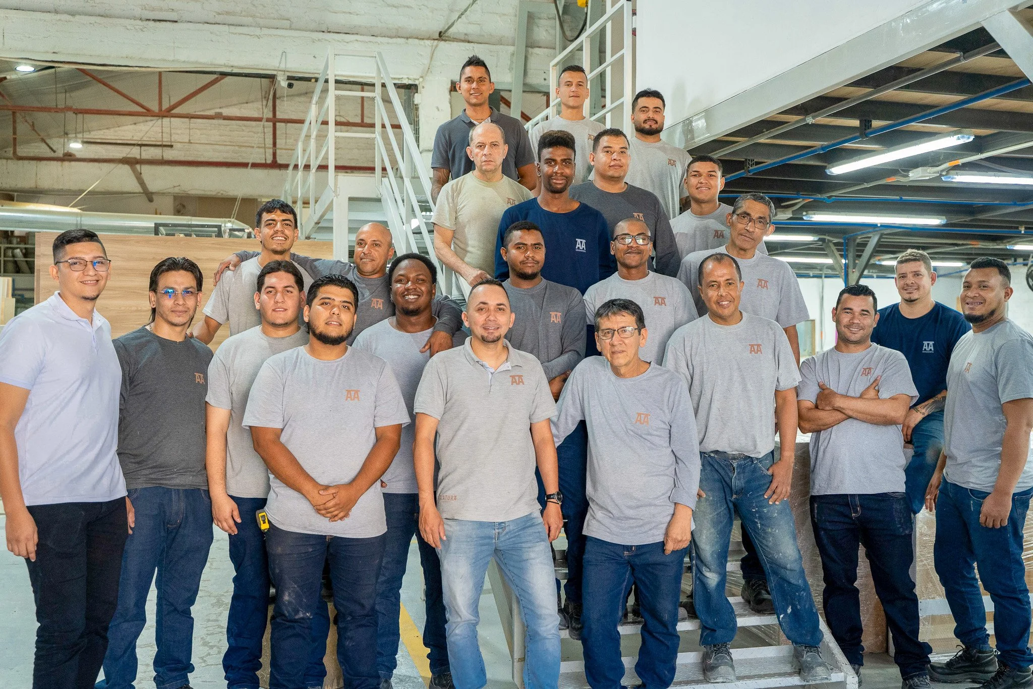 Grupo de personas en un entorno industrial, algunos llevan camisetas con logotipos y están de pie en diferentes niveles, posando para la foto.