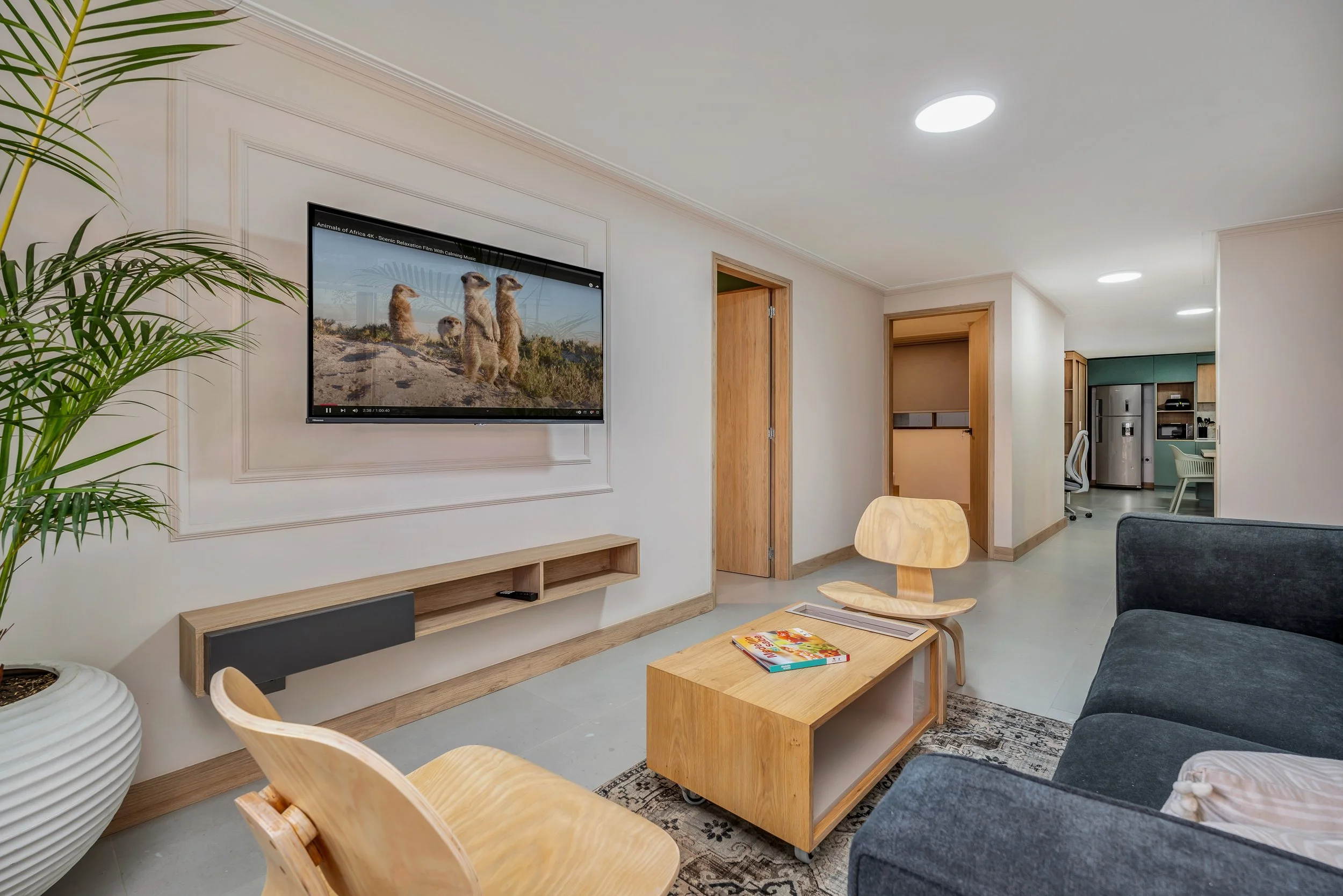 Sala de estar moderna con sofá gris, mesa de madera, televisión en la pared que muestra una escena de animales, plantas verdes en macetas y una cocina en el fondo.