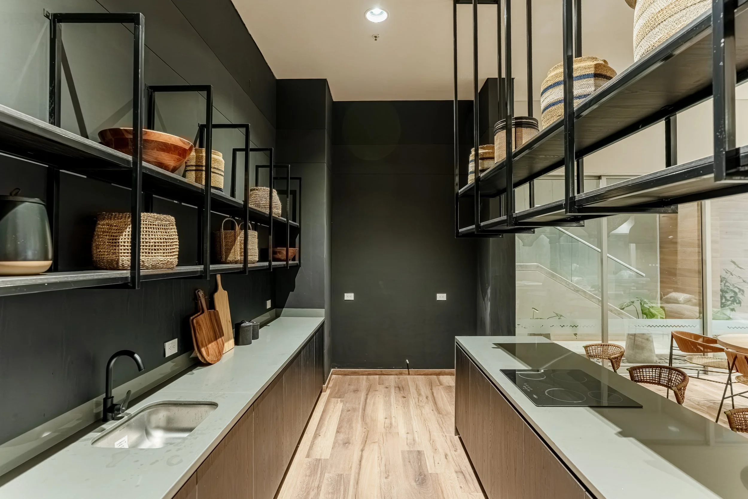 Cocina moderna con encimera de piedra, estanterías negras con decoraciones de cestas y vajilla, paredes en tonos oscuros y ventanas grandes que dejan entrar luz natural.