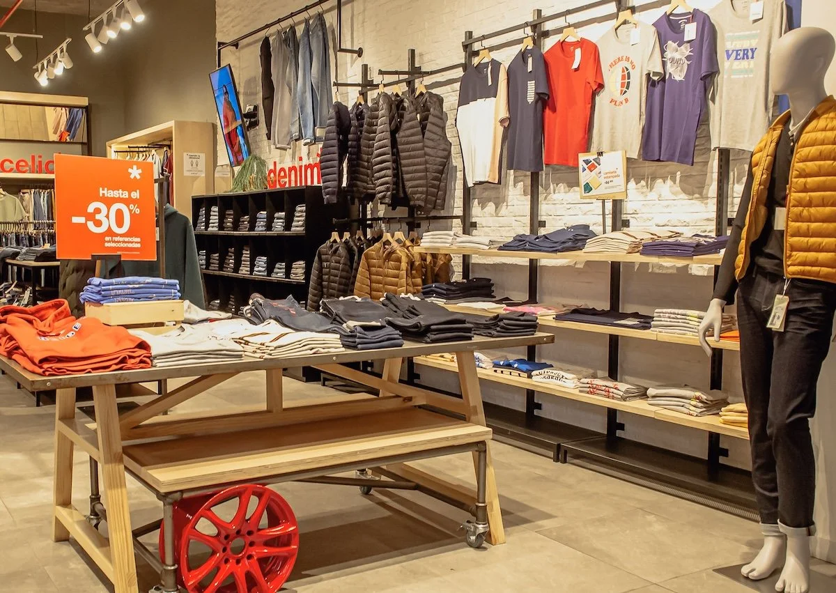 Se muestra una tienda de ropa con varias prendas y accesorios. En primer plano hay una mesa de madera con ropa doblada y un cartel que anuncia hasta un 30% de descuento. En el fondo hay estantes con camisetas, pantalones y chaquetas, y una pared con ropa colgada en perchas. A la derecha, un maniquí viste una chaqueta acolchada naranja y pantalones negros.