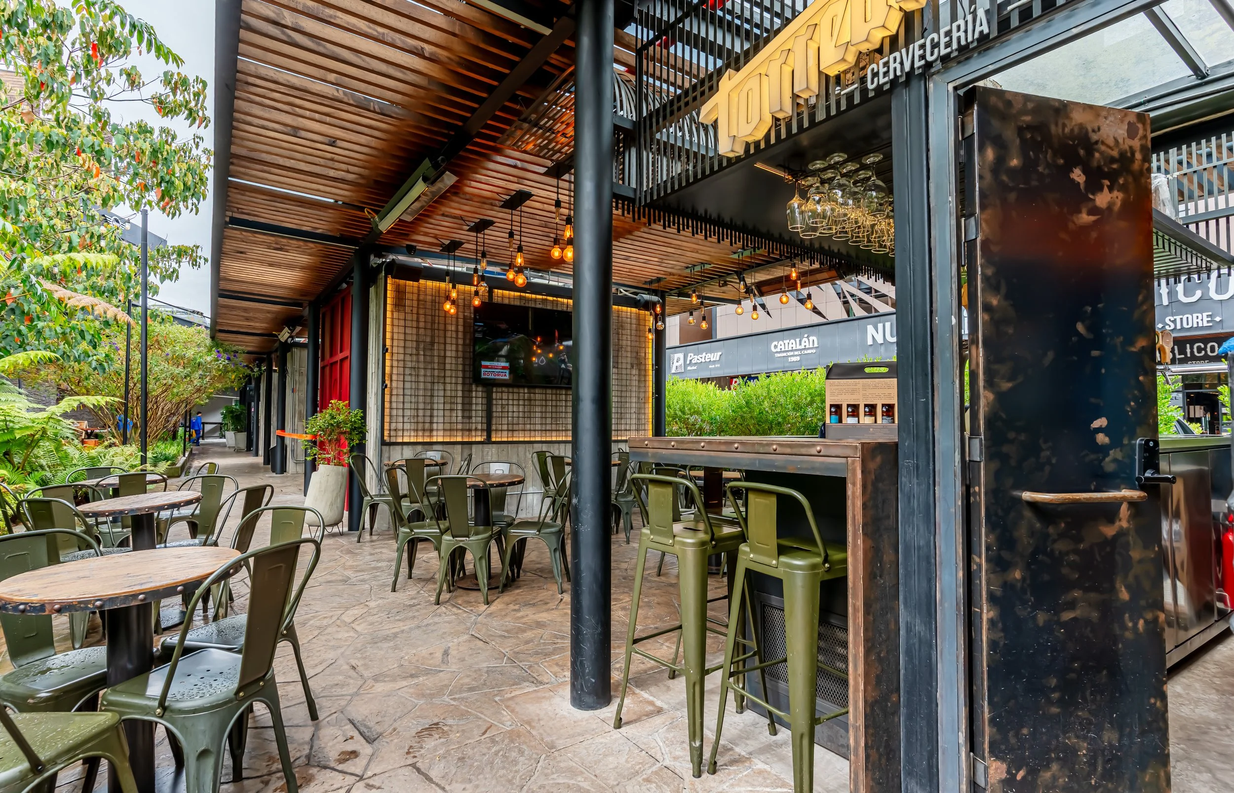 Área de descanso en una cafetería o restaurante con mesas de madera, sillas verdes, plantas decorativas, iluminación cálida con bombillas expuestas y un ambiente moderno de interior, con un toldo de madera en el techo y una vista parcial a tiendas vecinas.