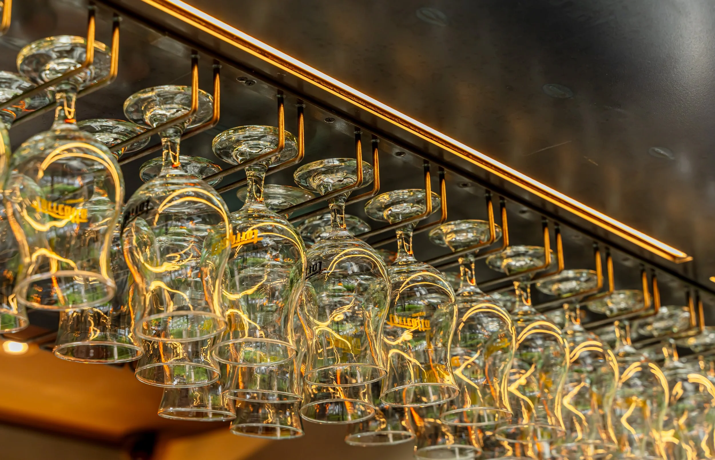 Vasos de vidrio colgados de un estante con soporte dorado, en un establecimiento, reflejando la iluminación cálida del lugar.