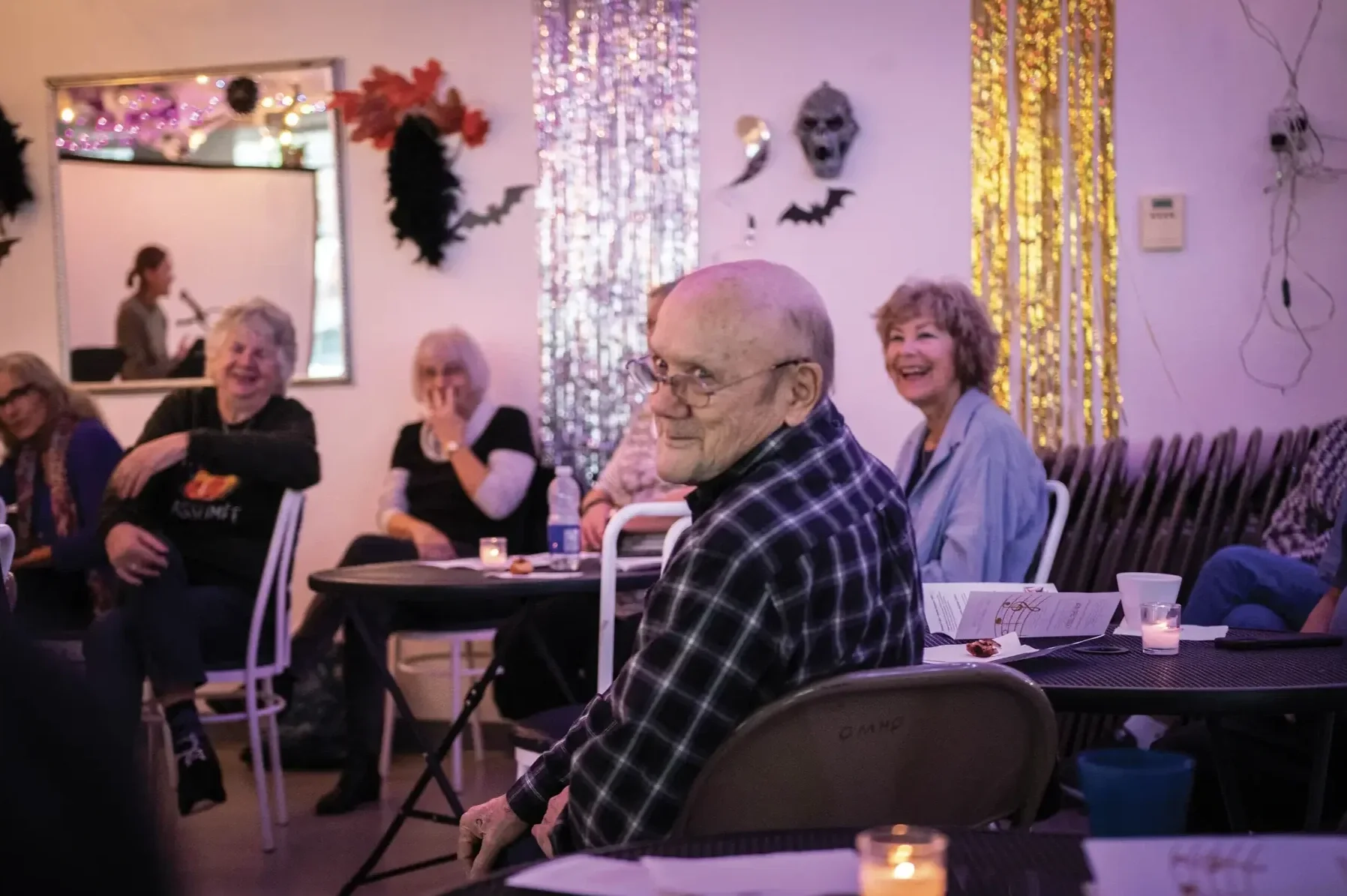 Un groupe de personnes âgées assises autour d'une table lors d'une fête, avec des décorations d'Halloween telles que des masques, des chauves-souris et des guirlandes ornementales, tout en riant et en discutant dans une ambiance festive et lumineuse.