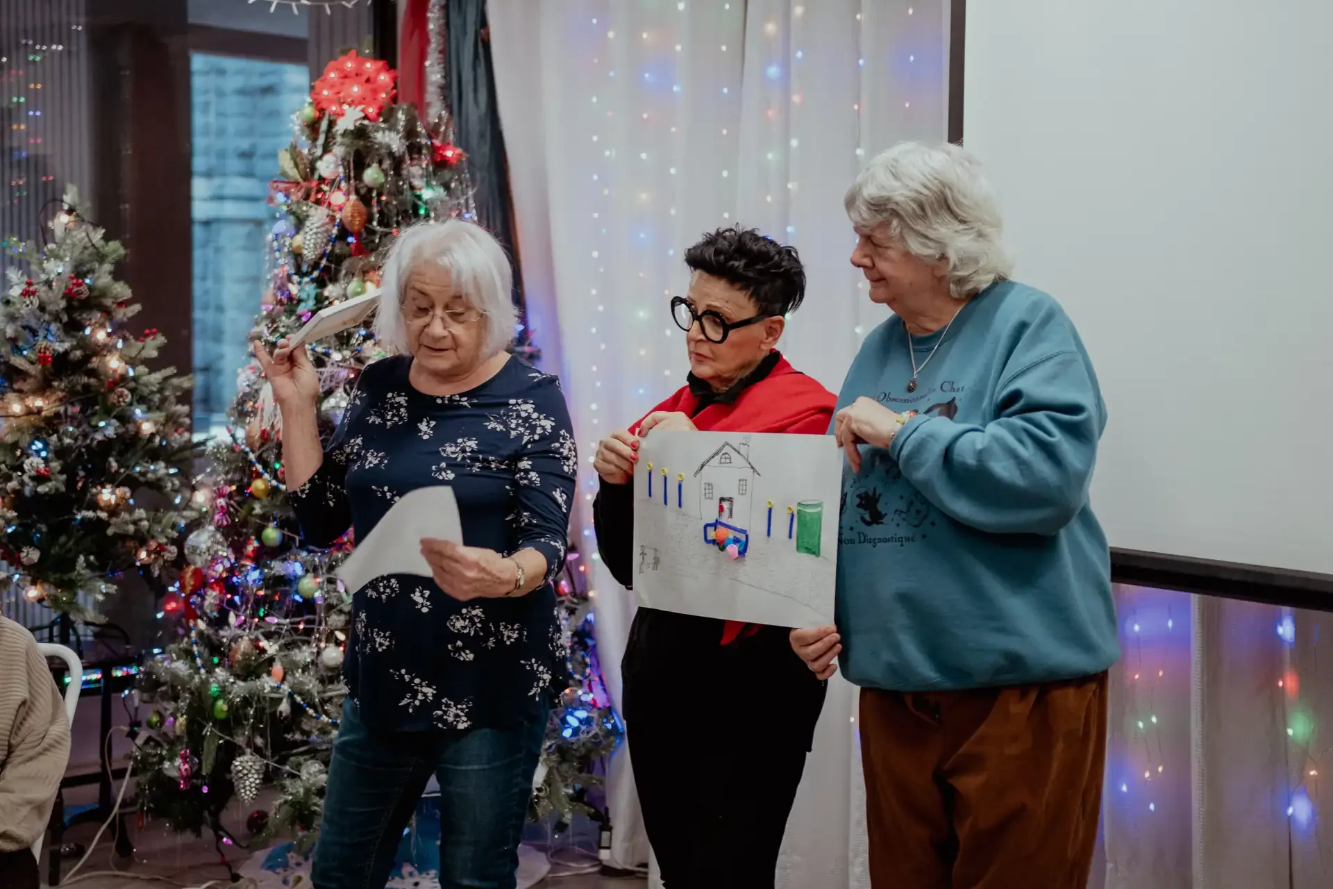 Trois personnes âgées participent à une célébration de Noël près d'un sapin décoré avec des boules et des guirlandes lumineuses. L'une d'elles tient une affiche avec un dessin d'une maison et des éléments de décoration.