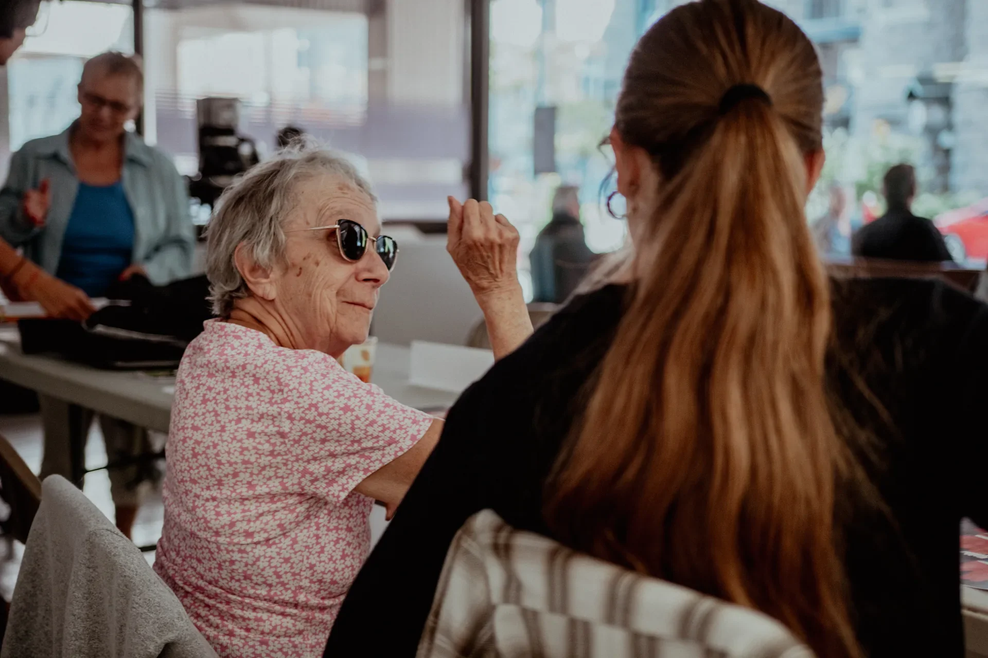 Une femme âgée portant des lunettes de soleil sourit en face d'une autre femme, dans un café ou un restaurant, avec d'autres personnes en arrière-plan.