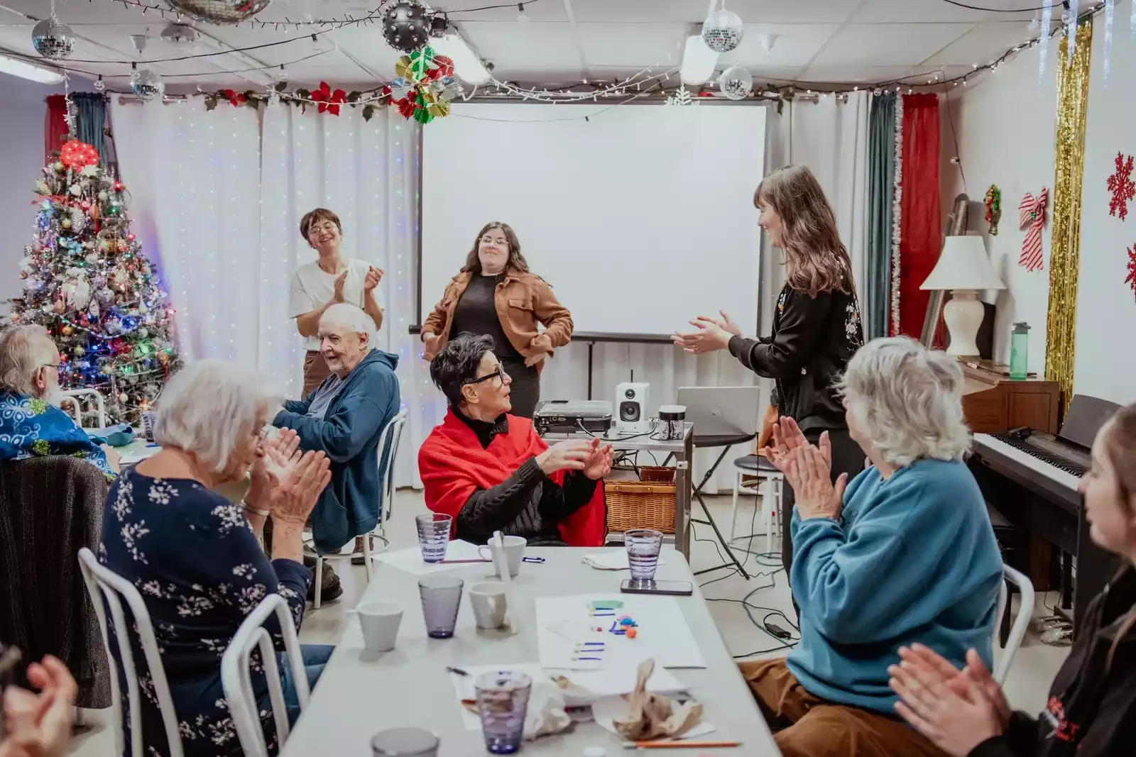 Groupe de personnes âgées assistant à une fête de Noël, avec un sapin décoré et des décorations festives, certains applaudissant ou souriant.