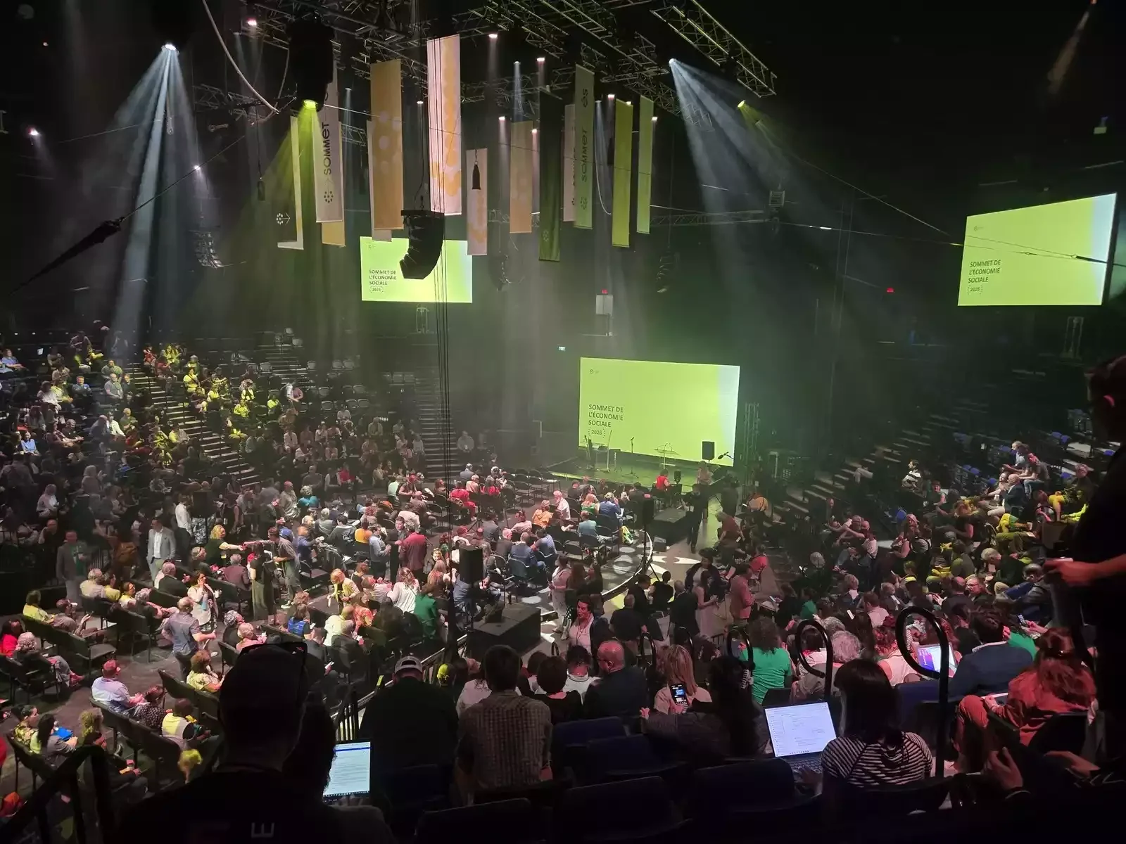 Une grande salle de conférence remplie de personnes assises, avec un grand écran vert affichant 'Sommet de l'économie sociale'. Il y a plusieurs écrans suspendus au plafond, des lumières et du matériel audiovisuel.