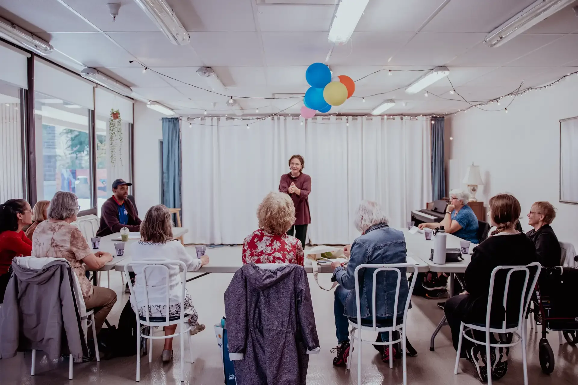 Une femme souriante parle devant un groupe de personnes assises autour d'une table, dans une pièce décorée avec des ballons colorés et des guirlandes lumineuses.