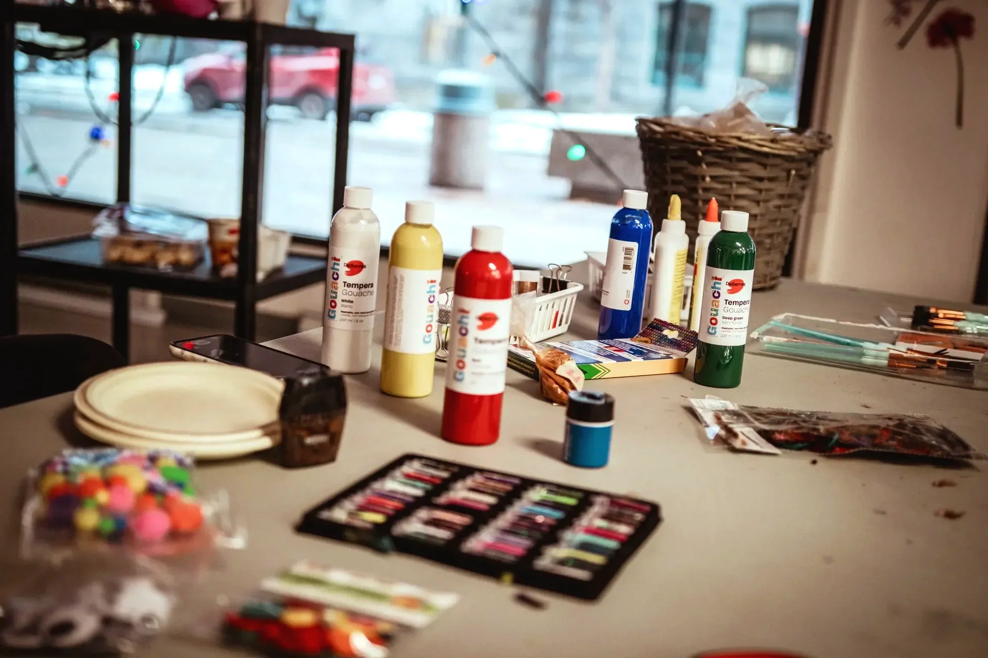 Table avec des tubes de peinture, pinceaux, et autres fournitures artistiques, près d'une grande fenêtre.