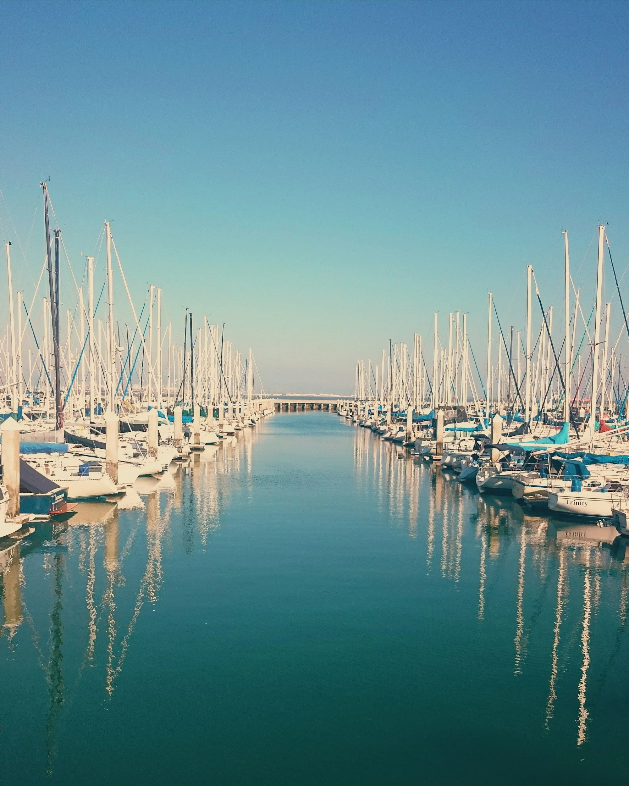 A marina with sailboats docked along both sides of a calm waterway, reflections of the boats visible on the water, under a clear blue sky.
