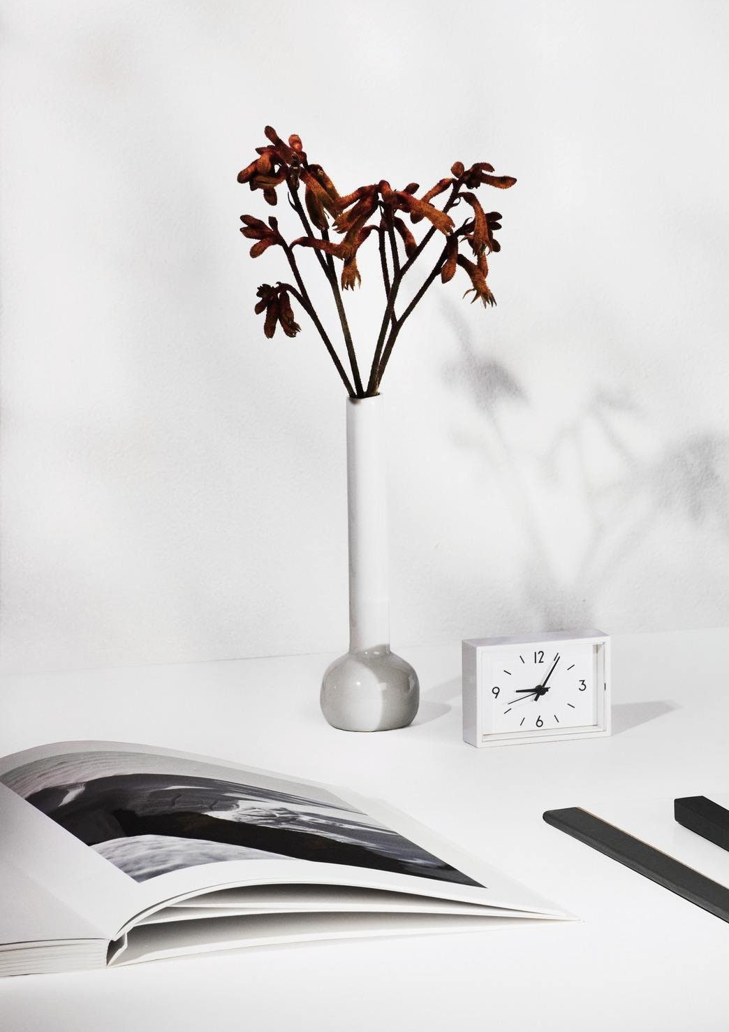 A minimalist white desk setup with a tall white vase holding dried brown flowers, a small white clock, an open book with black and white images, and two dark objects, all against a plain white wall.